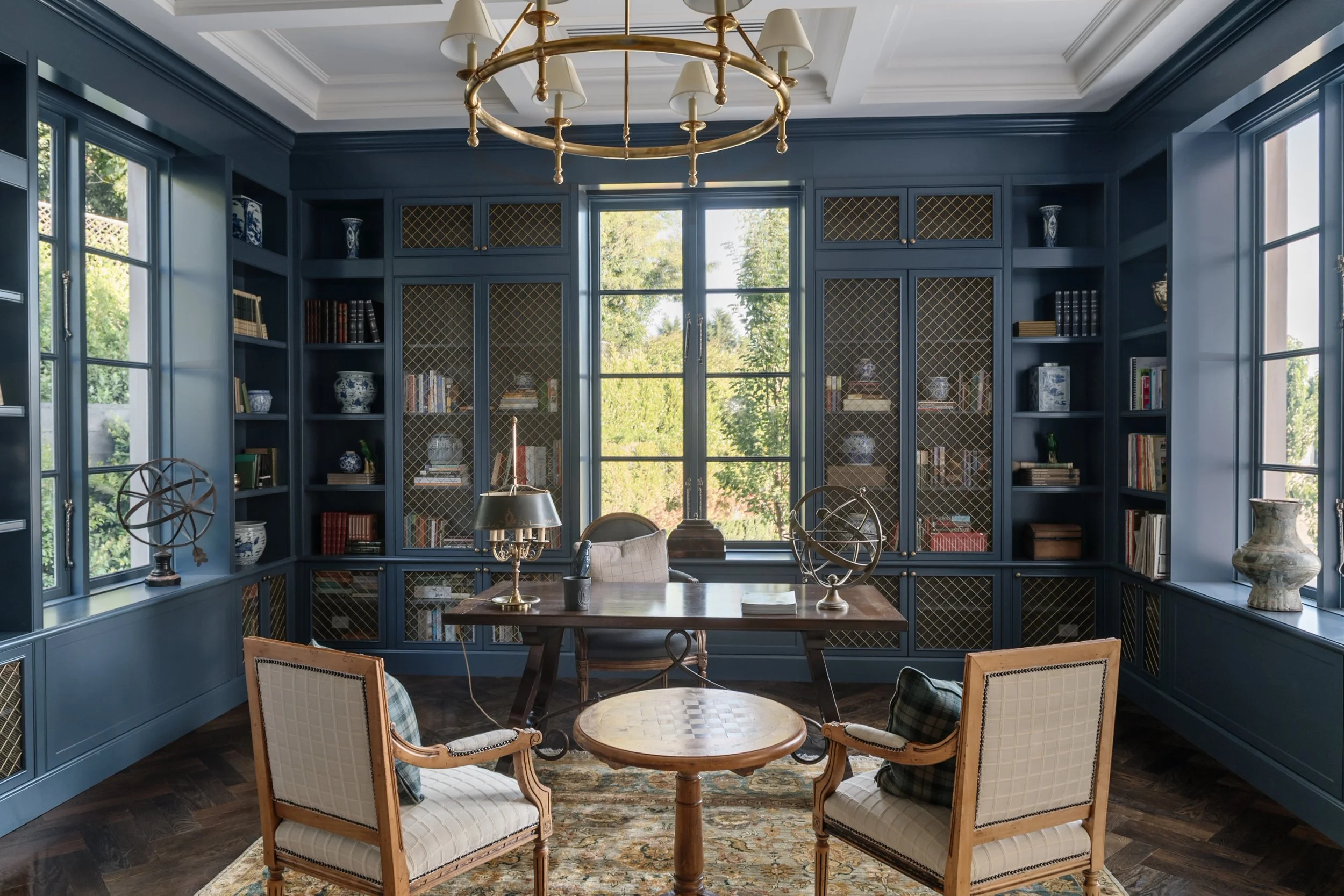 A home office with blue built-in bookshelves, a large window, a wooden desk, two vintage armchairs, and decorative items including globes, vases, and books.