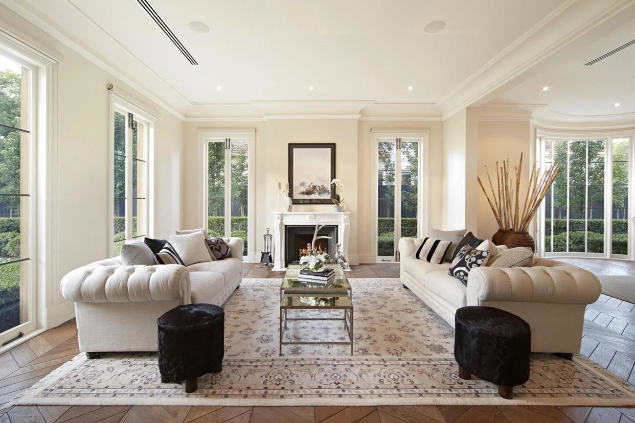 Bright living room with cream-colored sofas, black ottomans, a fireplace, large windows, and a patterned rug.