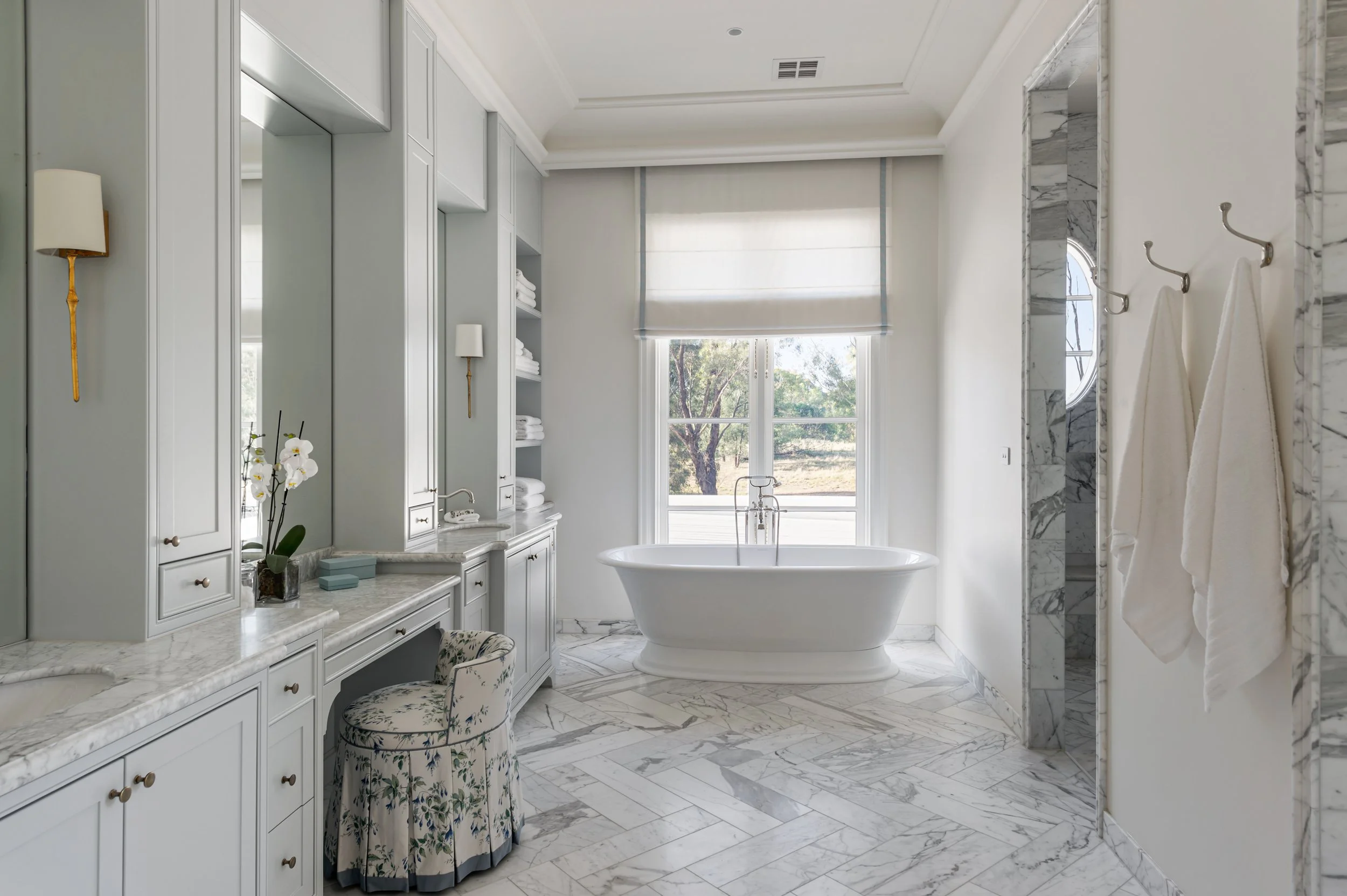 Bright bathroom with a freestanding bathtub in front of a large window, marble flooring and walls, built-in shelves with towels, a vanity with a floral upholstered stool, and towels hanging on hooks.