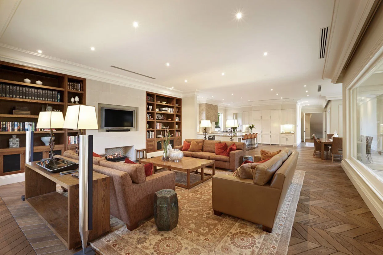Spacious living room with beige sofas, wooden coffee table, built-in bookshelves, a mounted flat-screen TV, and large windows, decorated in warm tones with hardwood floors and pendant lights.