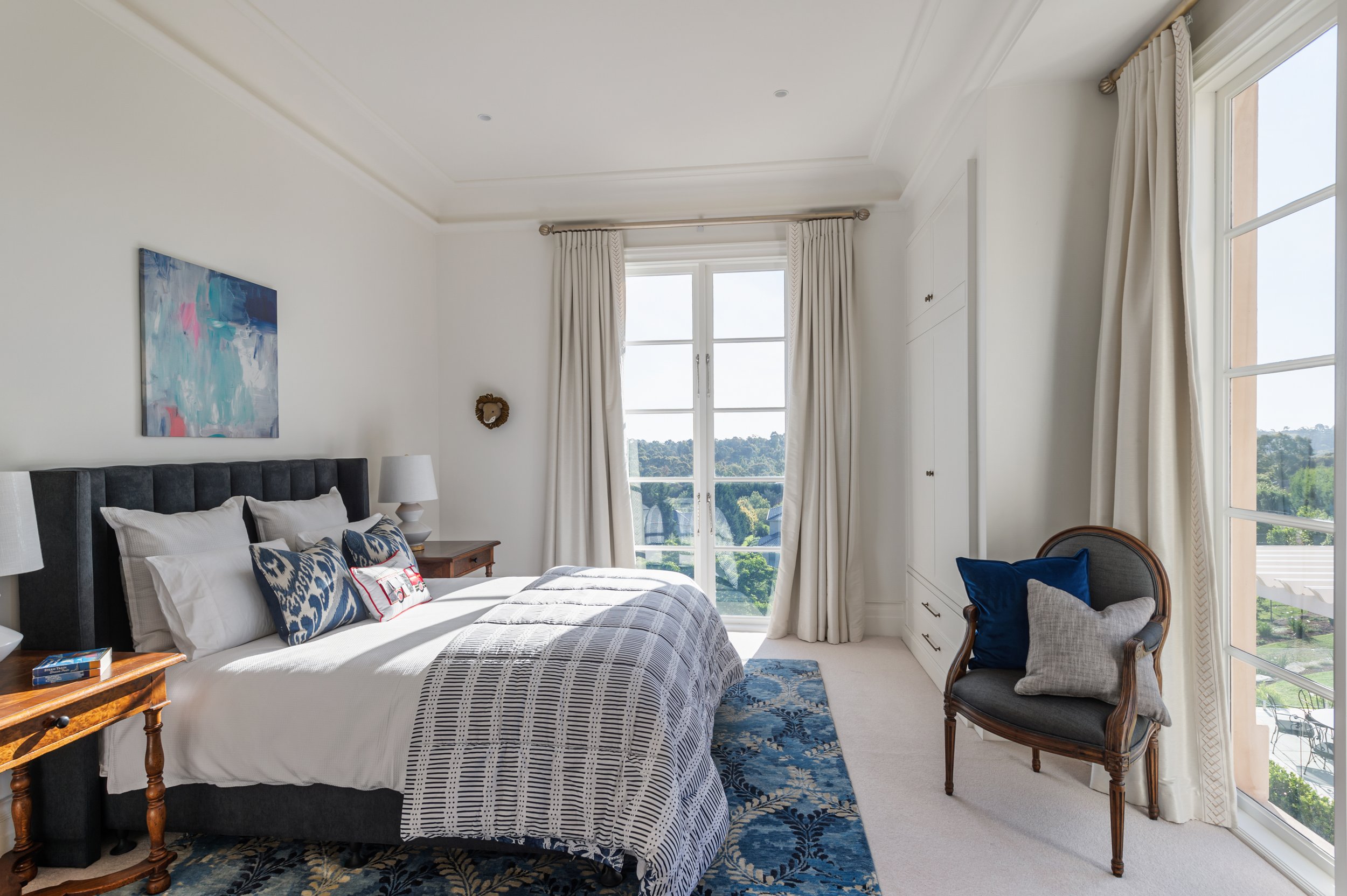 Bright bedroom featuring a large bed with white bedding and decorative pillows, nightstands with lamps, a blue patterned rug, a chair with two pillows, white curtains, and large windows showing an outdoor view.