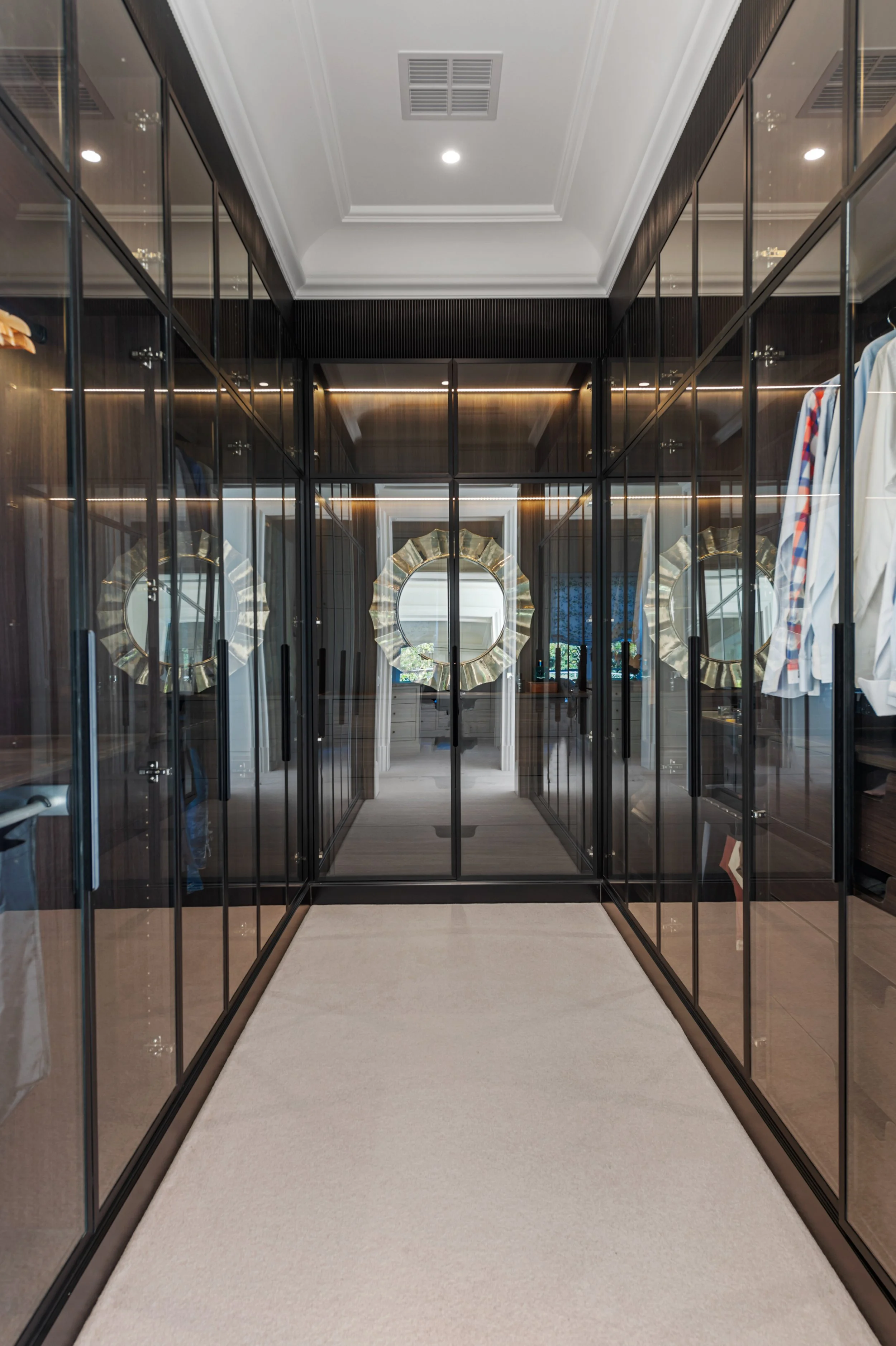 A walk-in closet with glass-fronted dark wood cabinets and clothing hanging, leading to a room with a large circular mirror decorated with a scalloped gold frame.