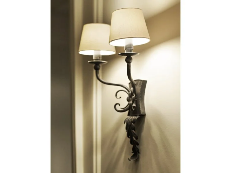 A decorative wall-mounted lamp with two beige lampshades and a black ornate metal arm and base, casting warm light on a wall.