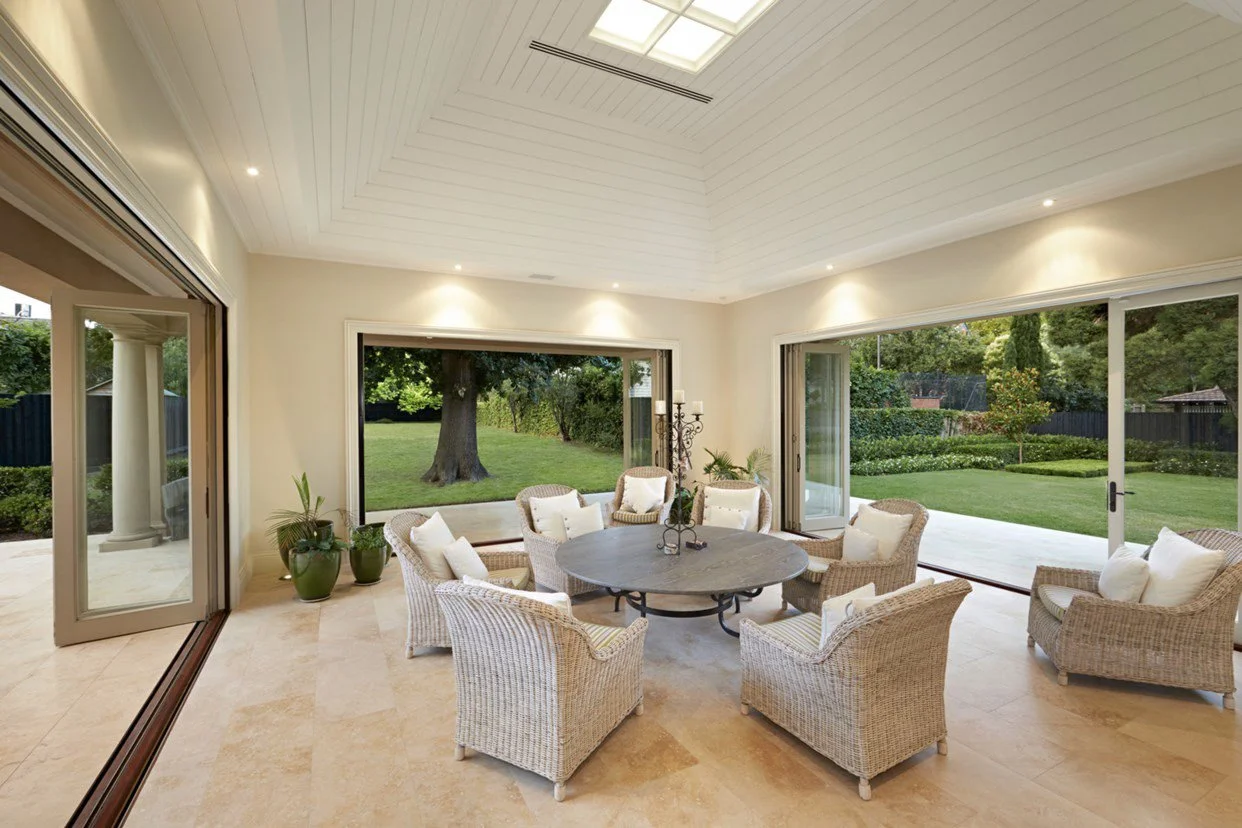 A bright indoor sunroom with sliding glass doors opening to a landscaped backyard with green lawn and trees. The room has a round table surrounded by eight wicker chairs with white pillows, and potted plants near the doors.
