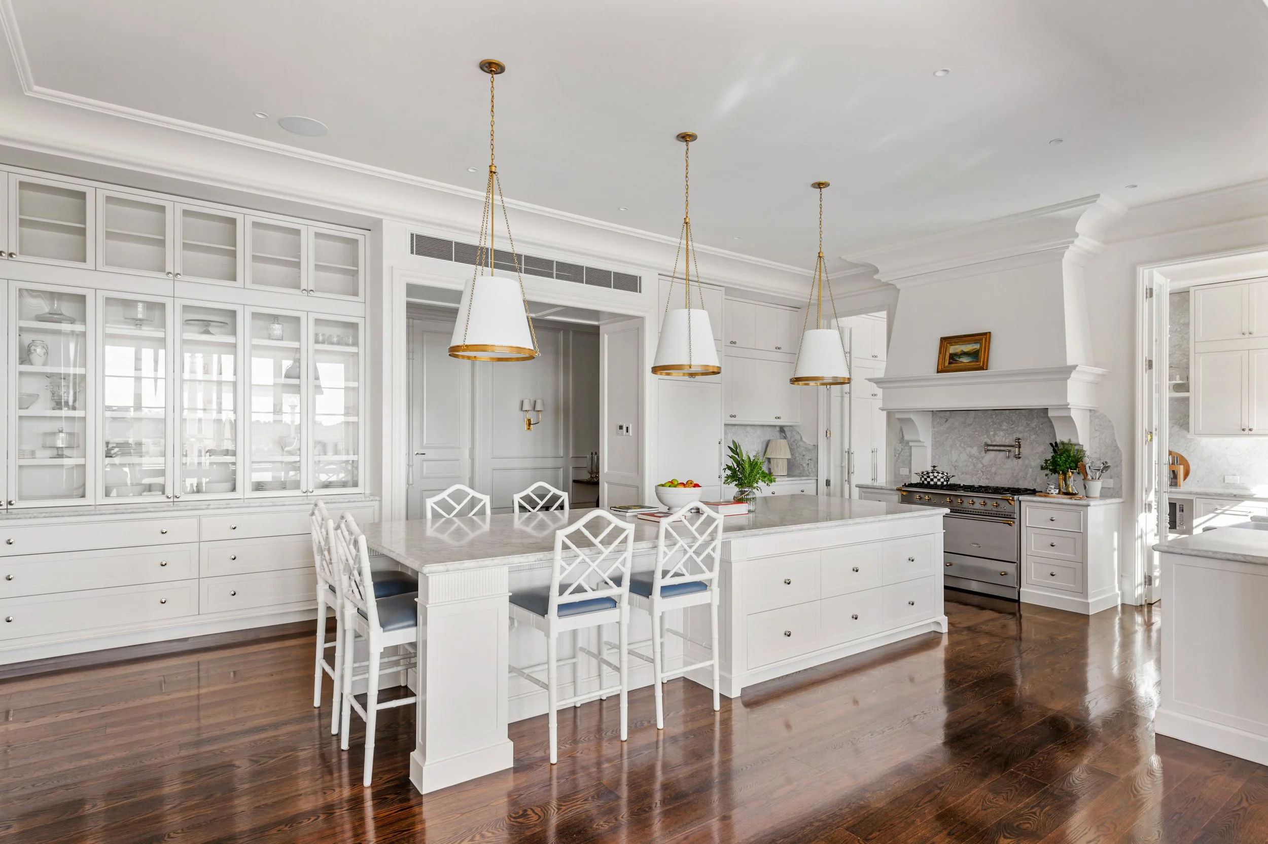 Bright white kitchen with large island, wooden flooring, glass-front cabinets, and a stove with a decorative mantle.