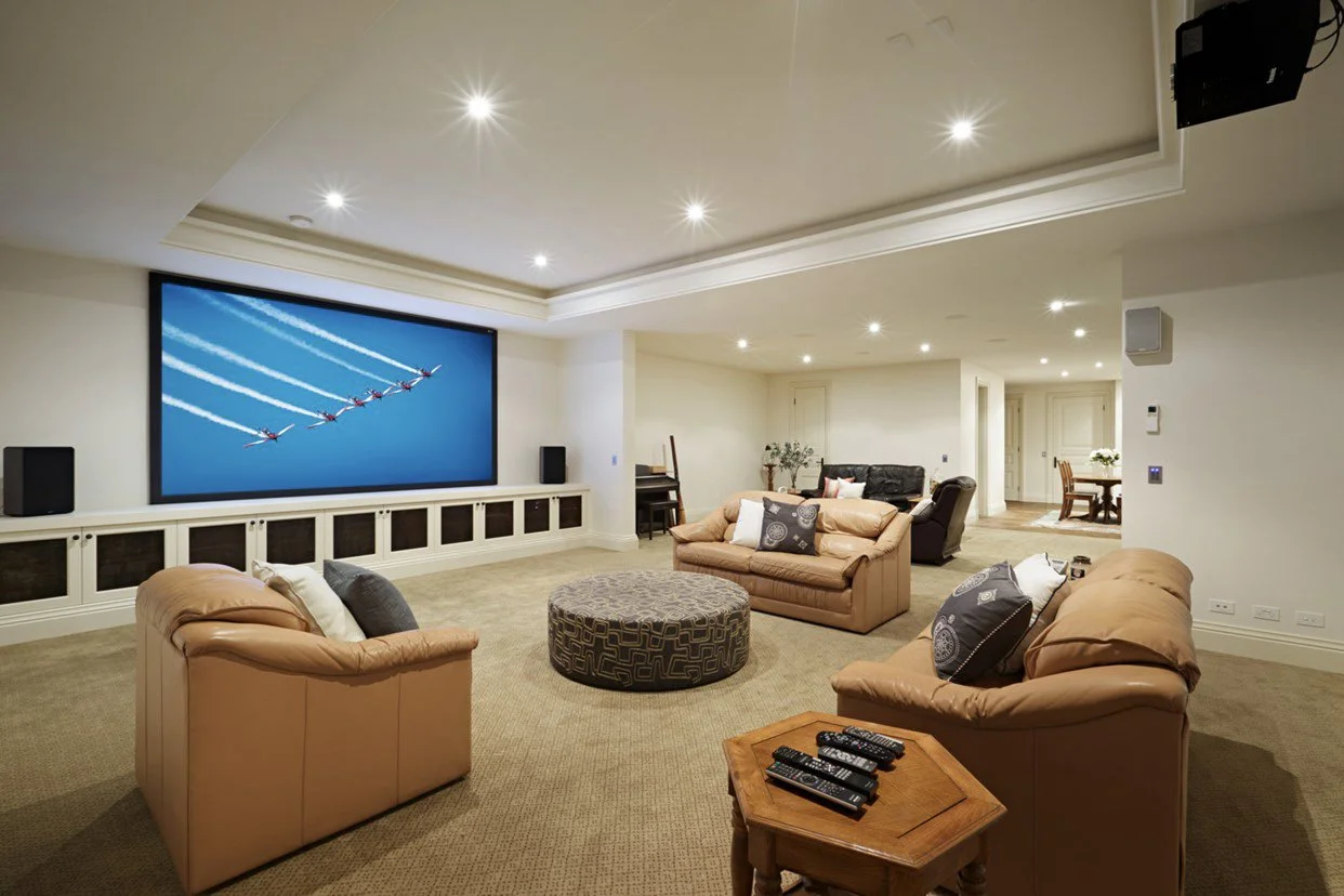 Spacious living room with beige sofas, a large flat-screen TV displaying an airplane formation, and a drum-shaped ottoman in the center. Multiple remote controls on a small table, carpeted floor, and built-in cabinetry beneath the TV. In the backgrou