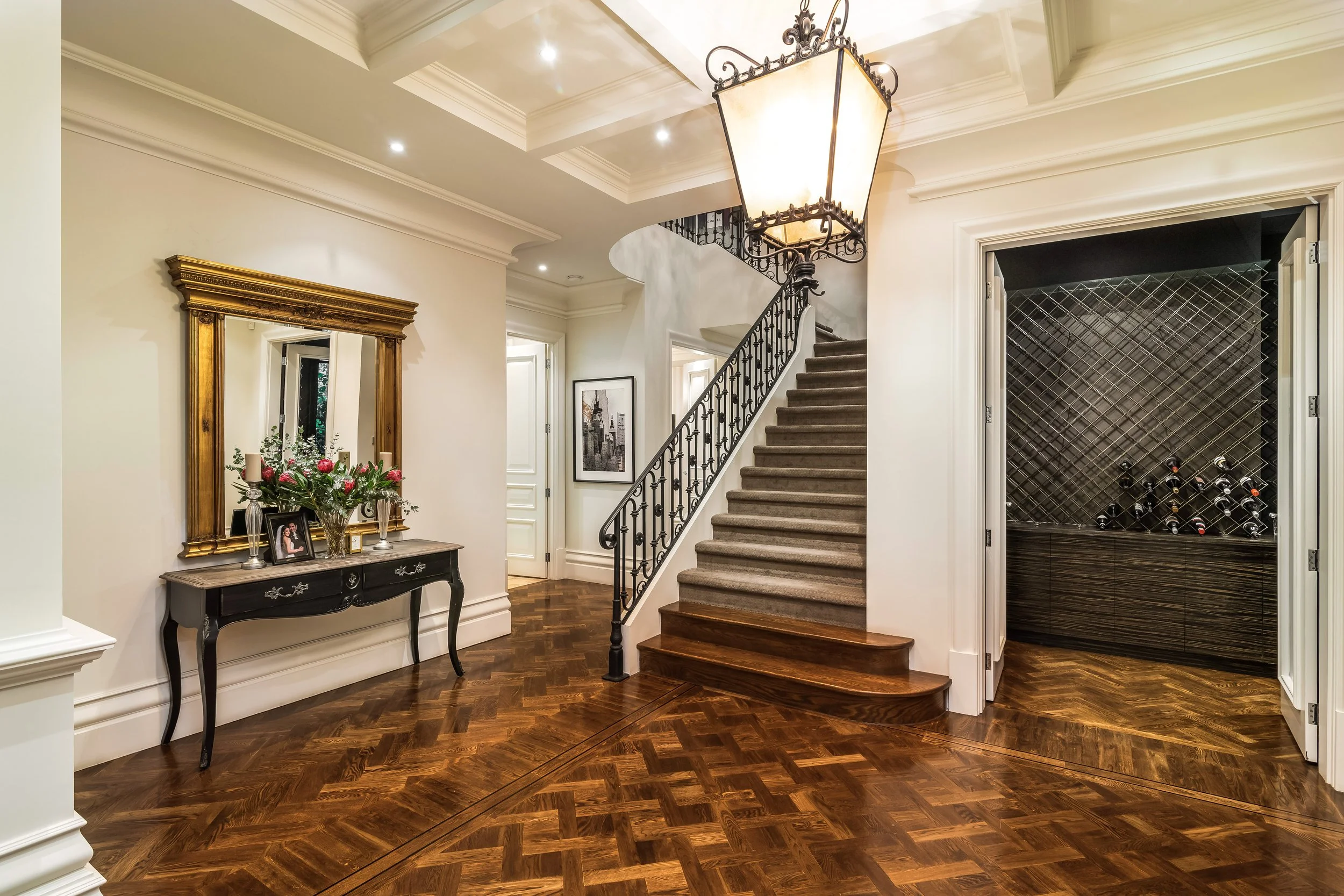 Elegant foyer with wooden parquet flooring, a staircase with carpeted steps and wrought iron railing, a large chandelier, a black console table with floral arrangement and framed photo, a large gilded mirror, and a wine storage area with bottles on r