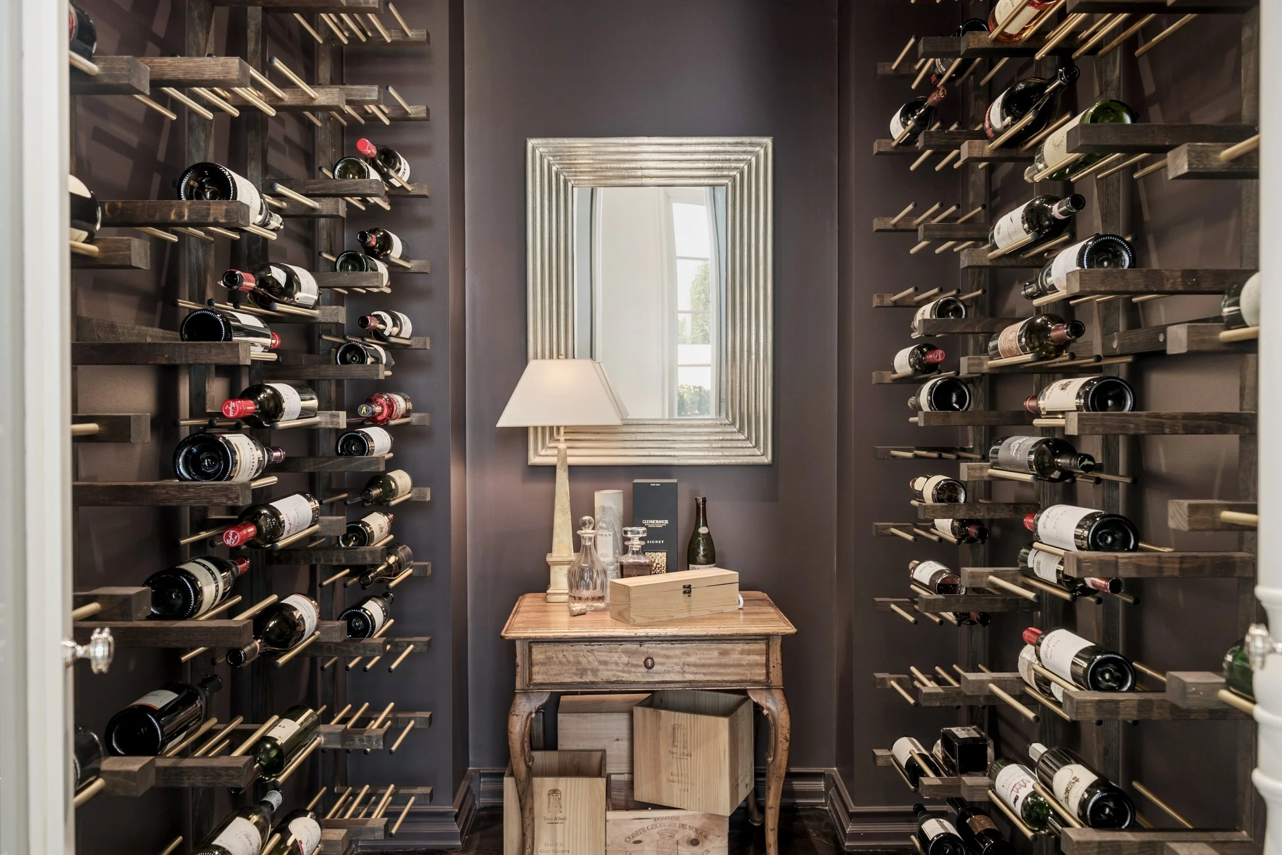 A cozy wine cellar with dark gray walls, flanked by wall-mounted wine racks filled with bottles. In the center, a small rustic wooden table holds glass bottles, a lamp, and other items. A large rectangular mirror with a silver frame hangs on the back