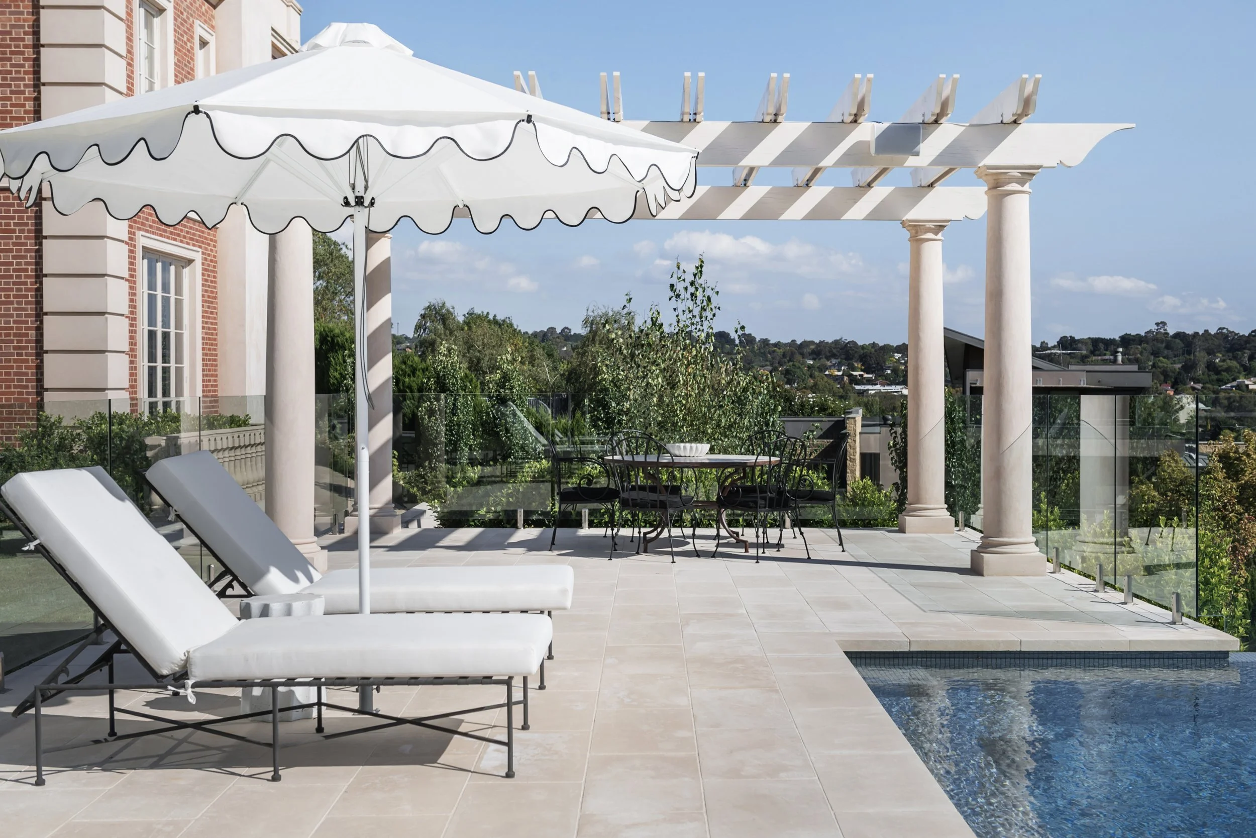 Luxury outdoor terrace with white lounge chairs, a large umbrella, a dining table with chairs, and a swimming pool, overlooking a cityscape with trees and houses under a blue sky.