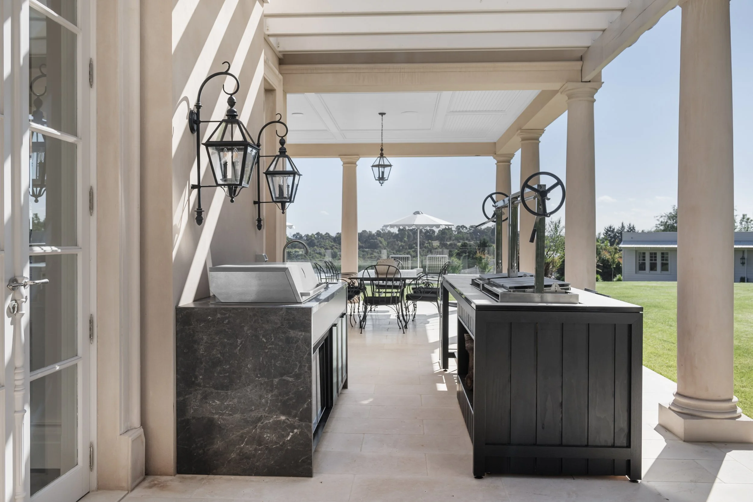 Outdoor kitchen and dining area with black countertops, metal chairs around a table, and a view of a backyard with grass and trees.