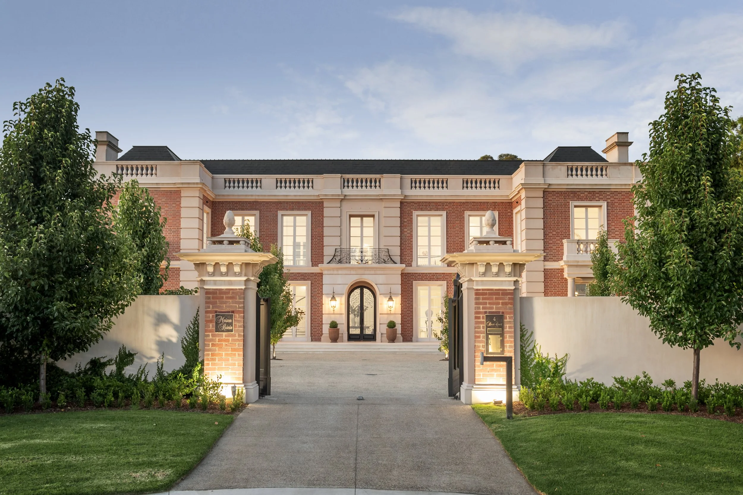 A grand, symmetrical brick mansion with white accents and tall windows, surrounded by a manicured lawn and trees, with a paved driveway leading to the front door.
