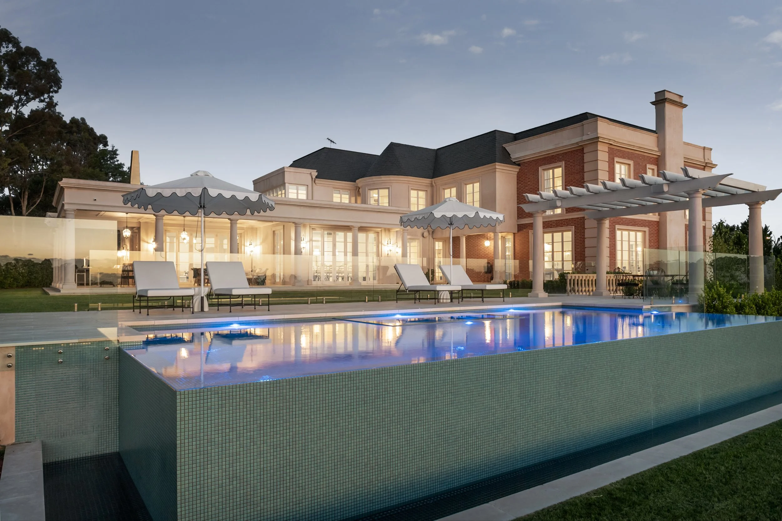 Elegant mansion with large windows, a swimming pool with blue lighting, outdoor lounge chairs with umbrellas, and a landscaped yard during twilight.