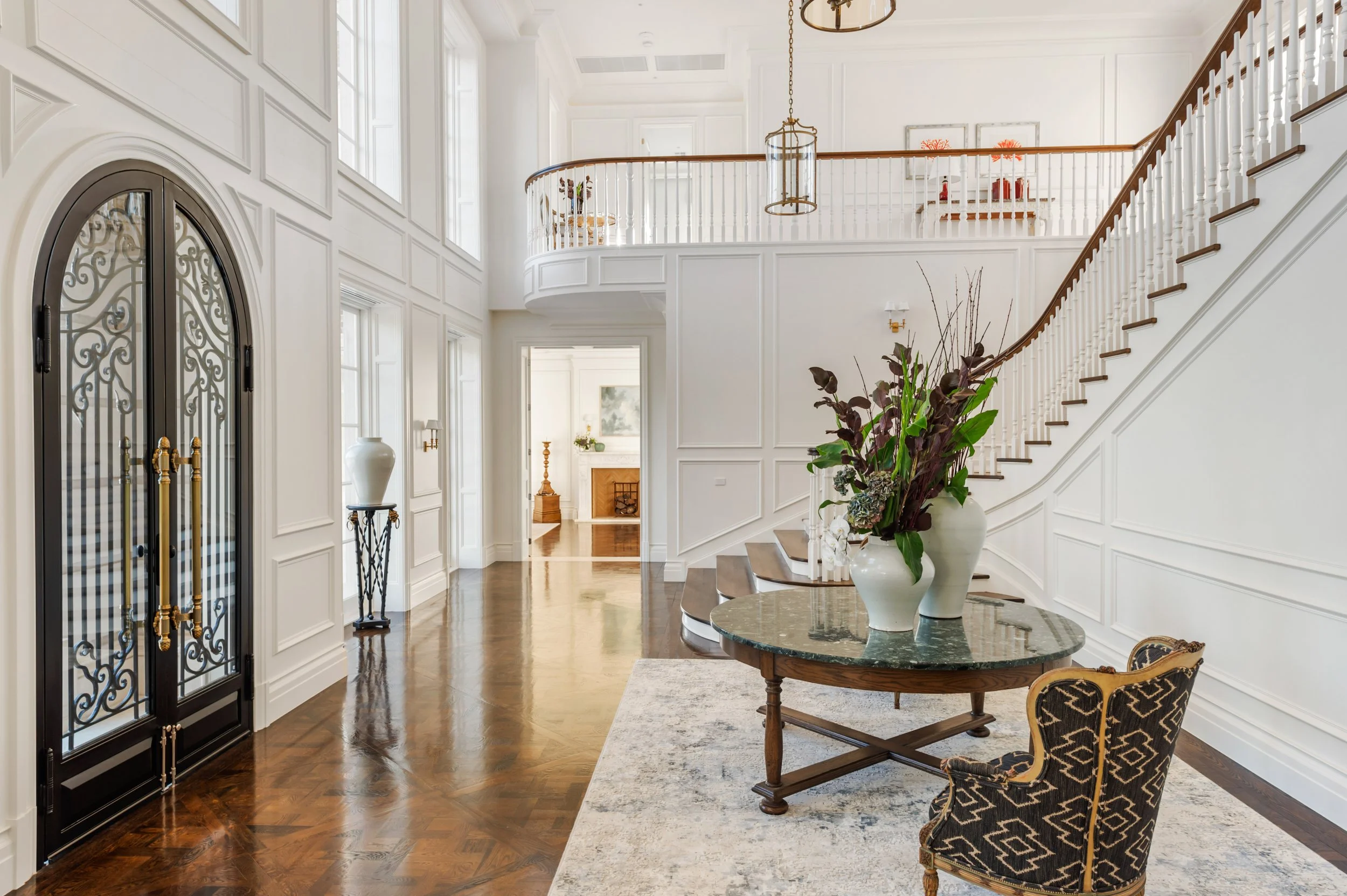 Spacious foyer with white walls and paneling, dark wood floors, a round table with large vases of purple and green foliage, an ornate black and gold chair, and a staircase with a curved wooden handrail and white spindles leading to a second floor.