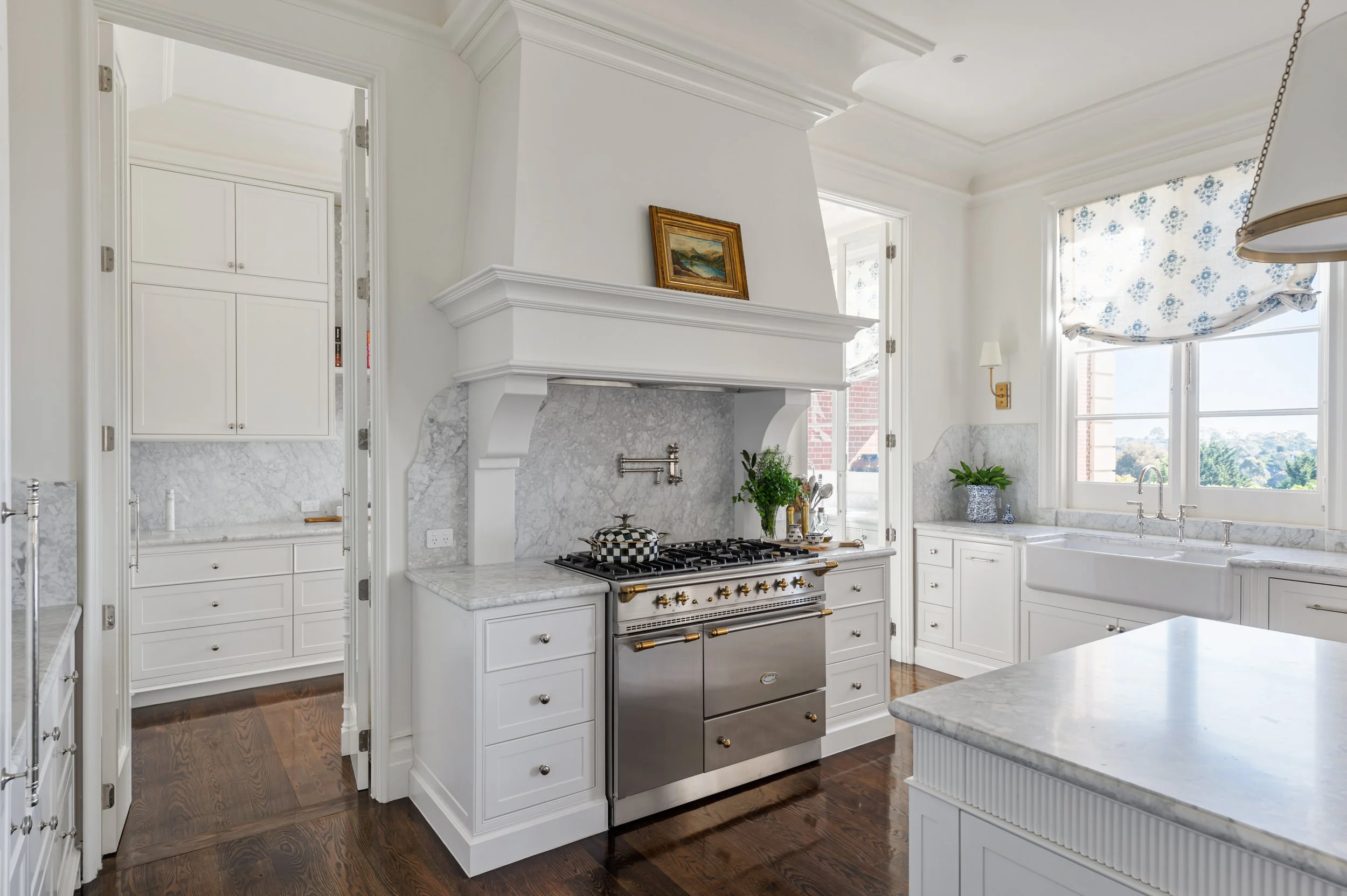Bright white kitchen with marble countertops, a stainless steel oven, and a window with a blue floral curtain.