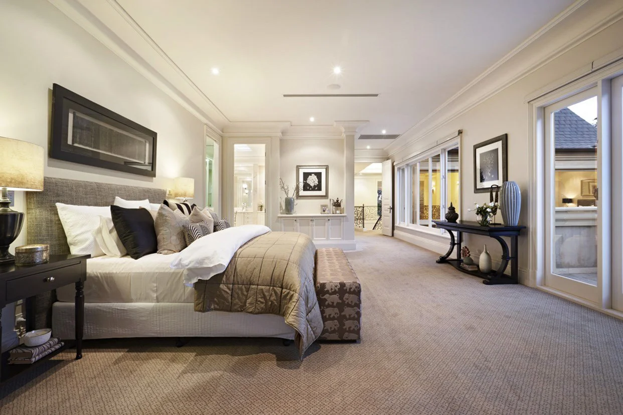 A spacious, well-lit bedroom with a large bed, multiple pillows, bedside tables with lamps, and decorative artwork on the walls. Large windows let in natural light.