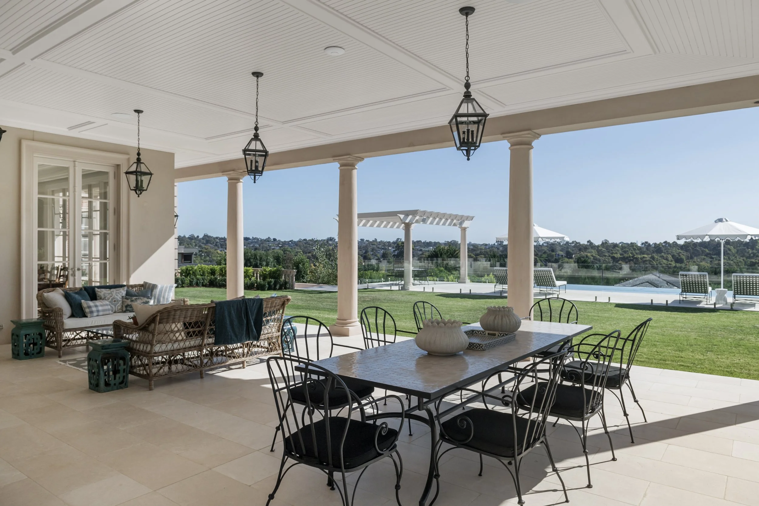 Covered outdoor patio area with black metal dining table and chairs, wicker sofa with cushions, and decorative planters on the table, overlooking a grassy backyard with umbrellas and lounge chairs, under a clear blue sky.