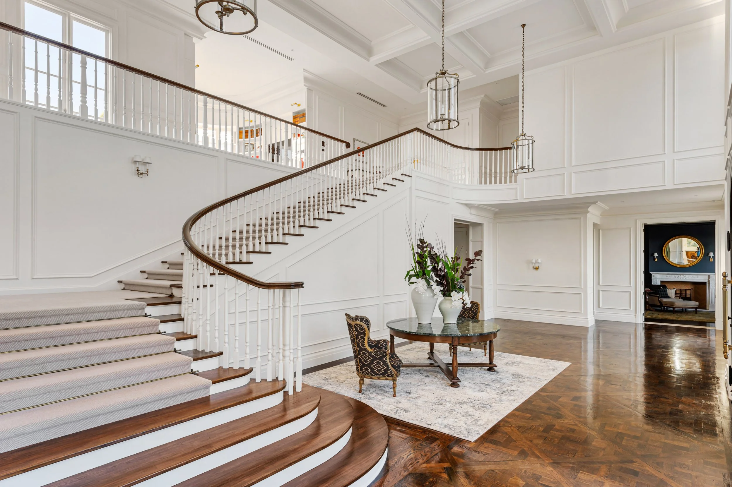 Elegant spacious foyer with a grand curved staircase, white paneled walls, dark hardwood floors, a round table with floral vases, and an adjacent sitting area with a fireplace and mirror.