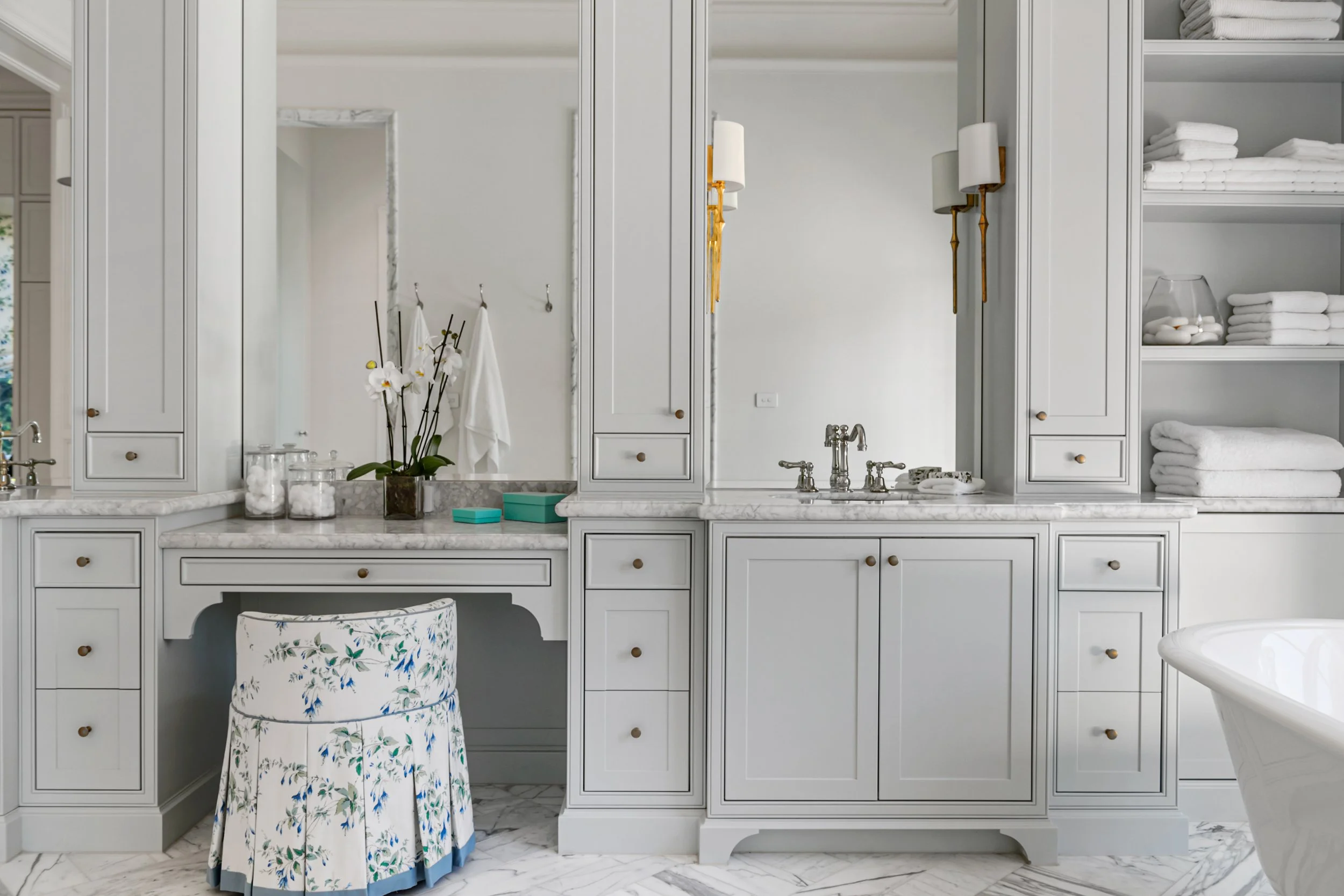 A luxurious white bathroom with marble countertops, a large framed mirror, wall-mounted light fixtures, and built-in shelves holding folded towels and toiletries.