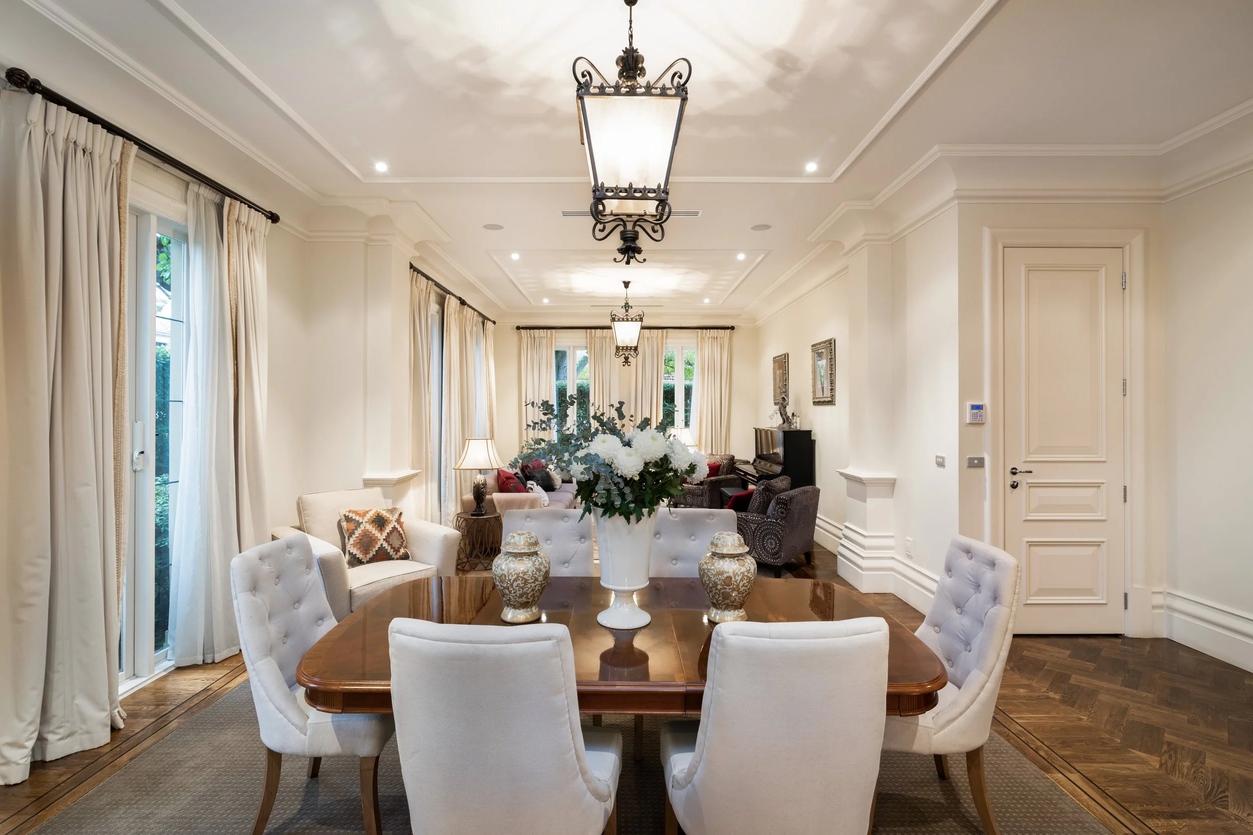 Elegant living and dining room with cream-colored walls, dark hardwood floors, large windows with cream curtains, central chandelier, and a round dining table with white upholstered chairs, a large floral centerpiece, and decorative vases.
