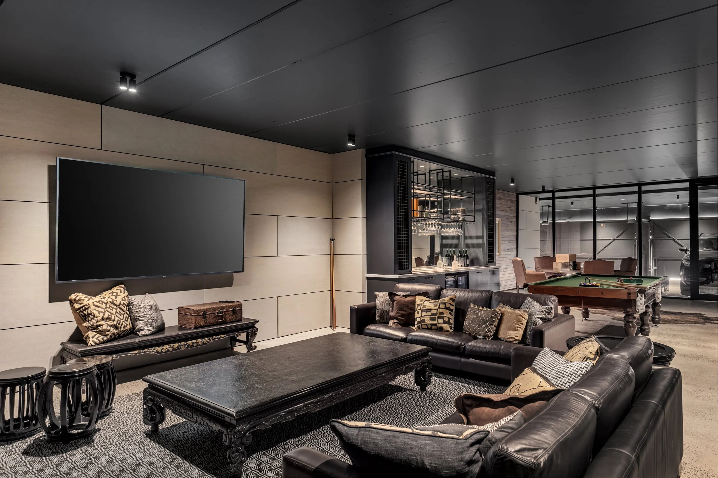 Modern basement with black leather sofas, a large flat-screen TV, a black ornate coffee table, patterned pillows, a pool table, and a bar area, with glass walls and dark ceiling lights.