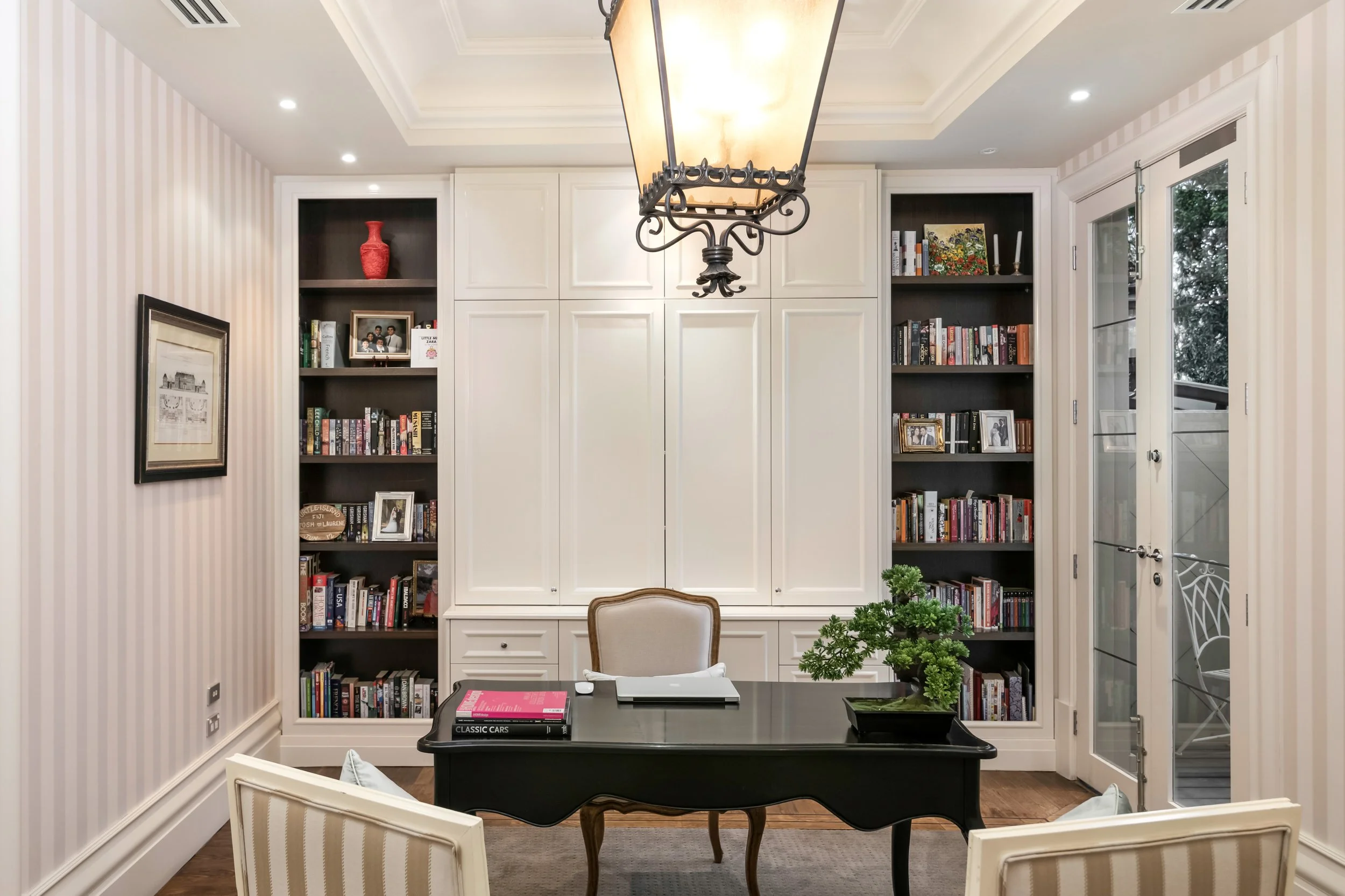 Home office with dark wood desk, beige chair, built-in white cabinets, black bookshelves filled with books and framed photos, potted plant on desk, wall art, and glass-paneled door leading to outdoor deck.