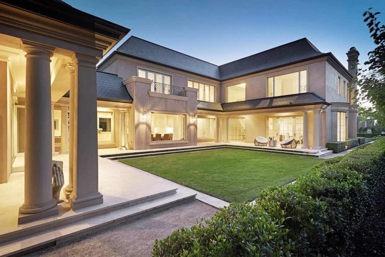 A large, elegant two-story house with bright interior lighting, a well-maintained lawn, and patio furniture during dusk.