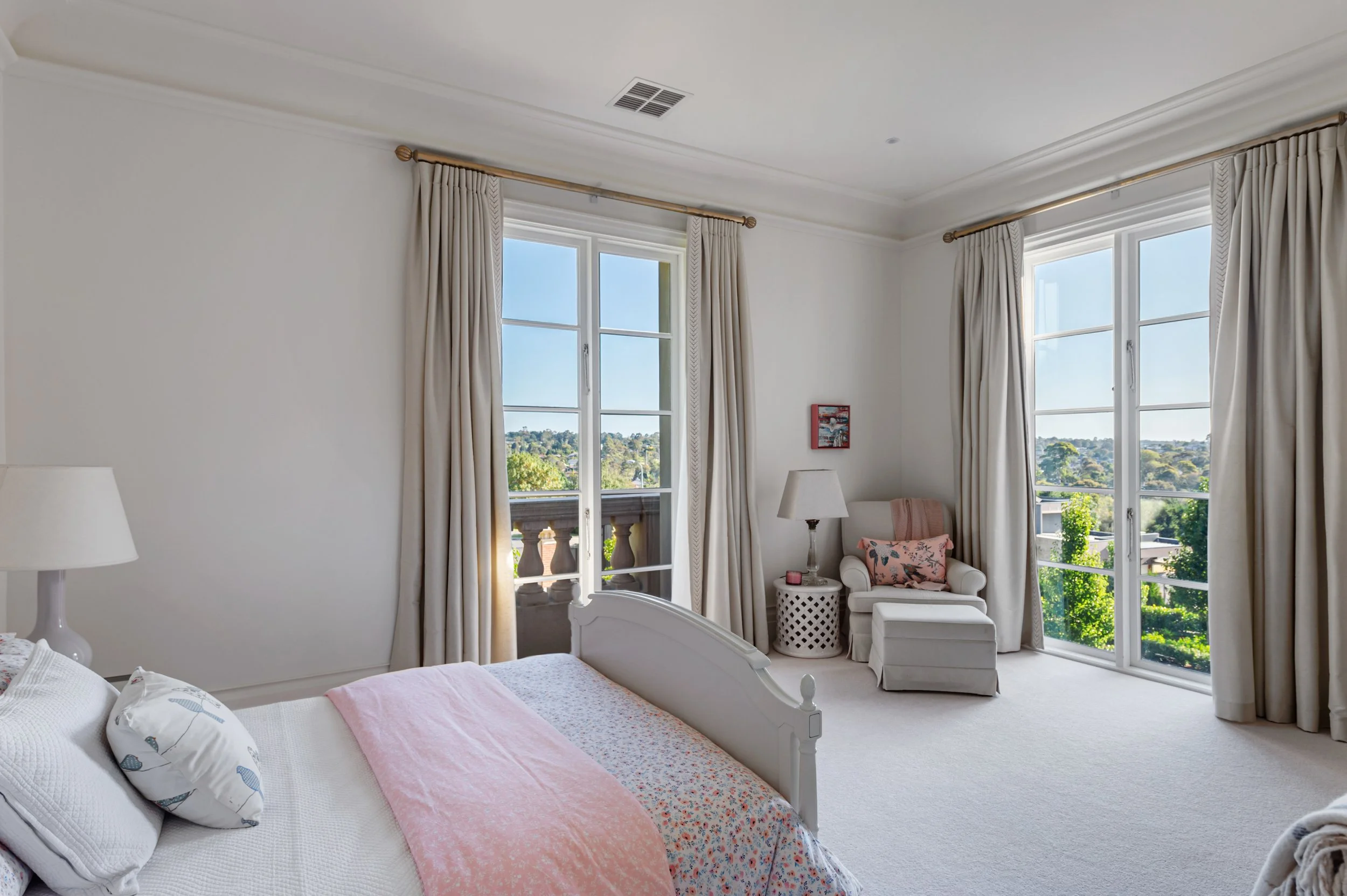 Bright bedroom with white walls, large windows with beige curtains, a white bed with pink and floral bedding, a white bedside table with a lamp, and a cozy seating area with an armchair, pink pillow, and view of green trees outside.