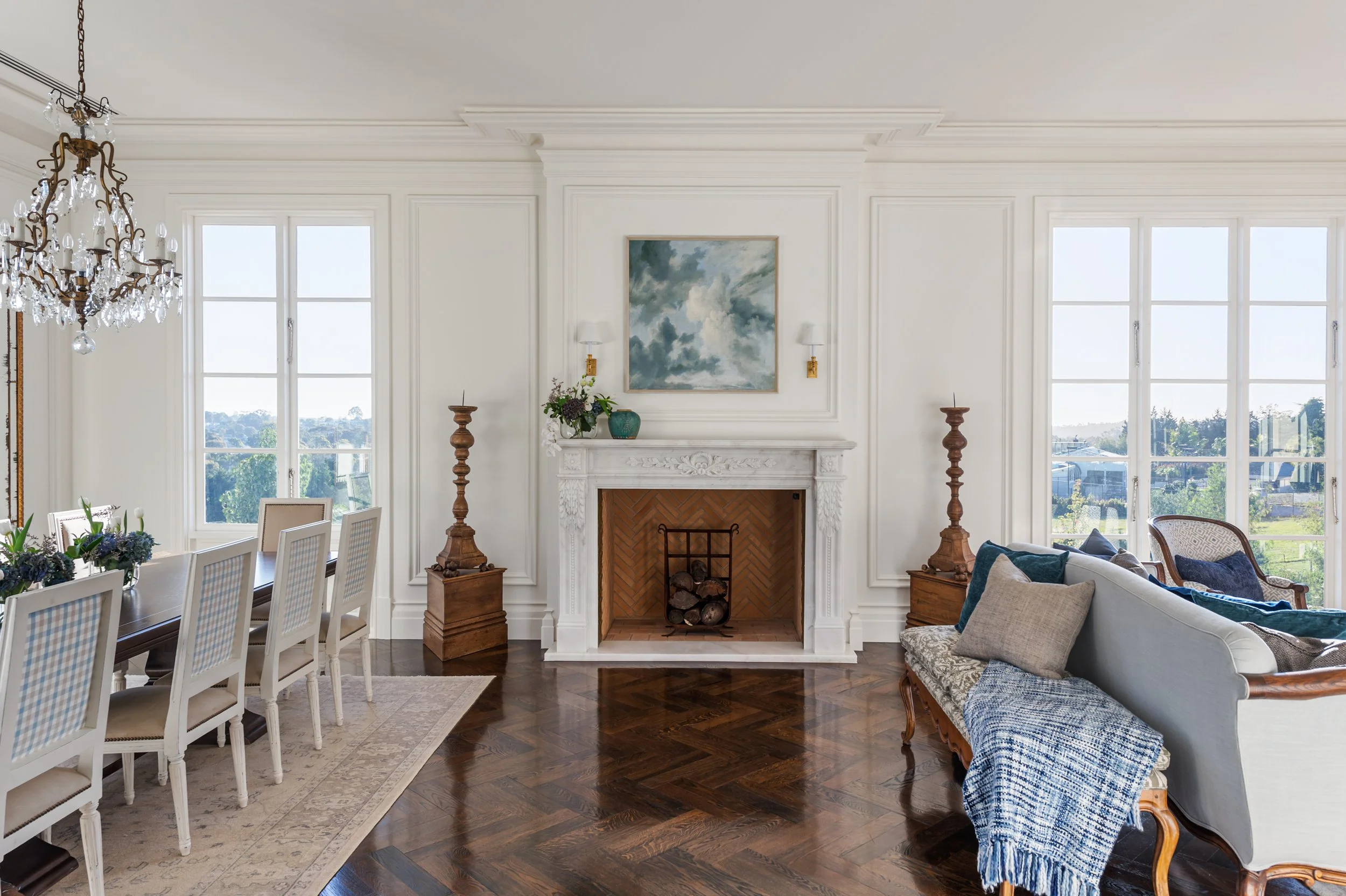 Living room with a white ornate fireplace, built-in windows on either side, a chandelier, a dining table with chairs, and a light blue sofa with cushions.