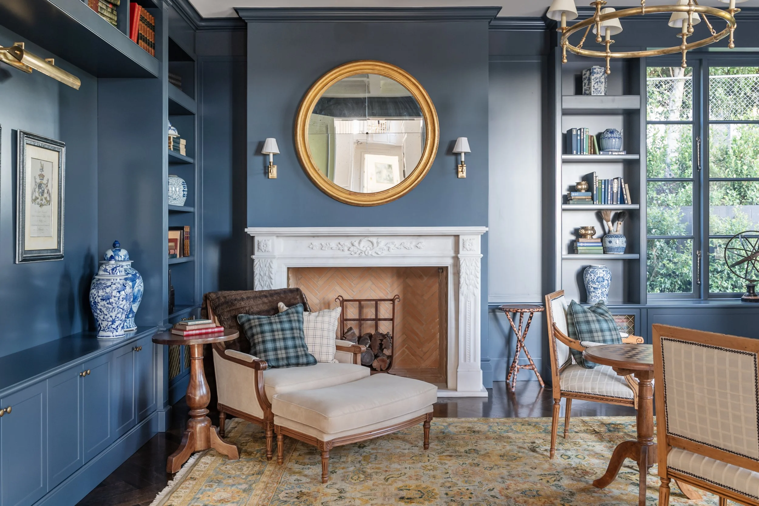 A cozy living room featuring dark blue walls, a white ornate fireplace, a large golden-framed round mirror above the fireplace, two small white lamps mounted on the wall, a brown upholstered armchair with a plaid pillow, a cream chaise lounge with pl