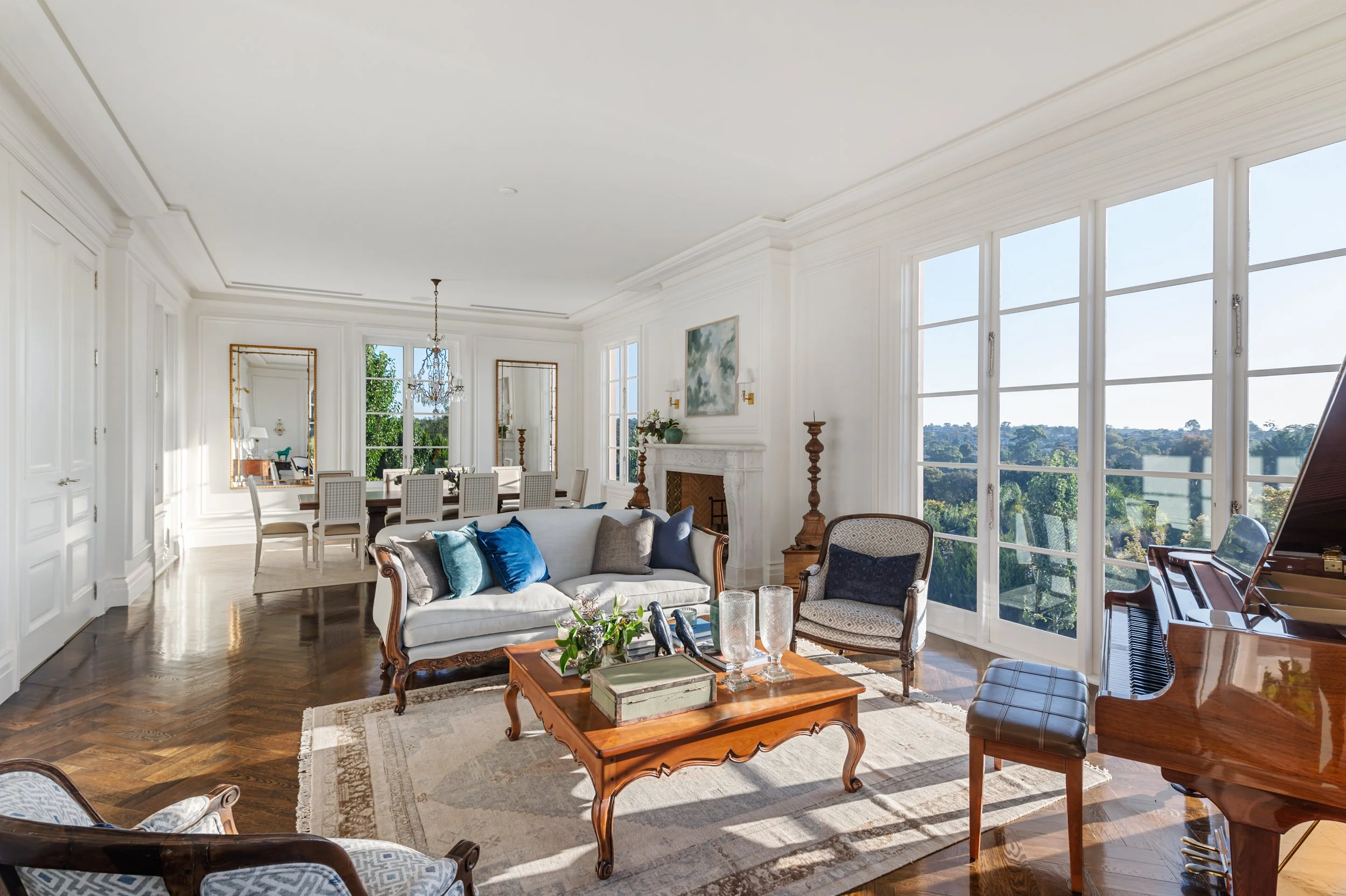 Spacious living room with white walls, large windows, hardwood floors, elegant furniture, a grand piano, and a fireplace with a framed painting above it.