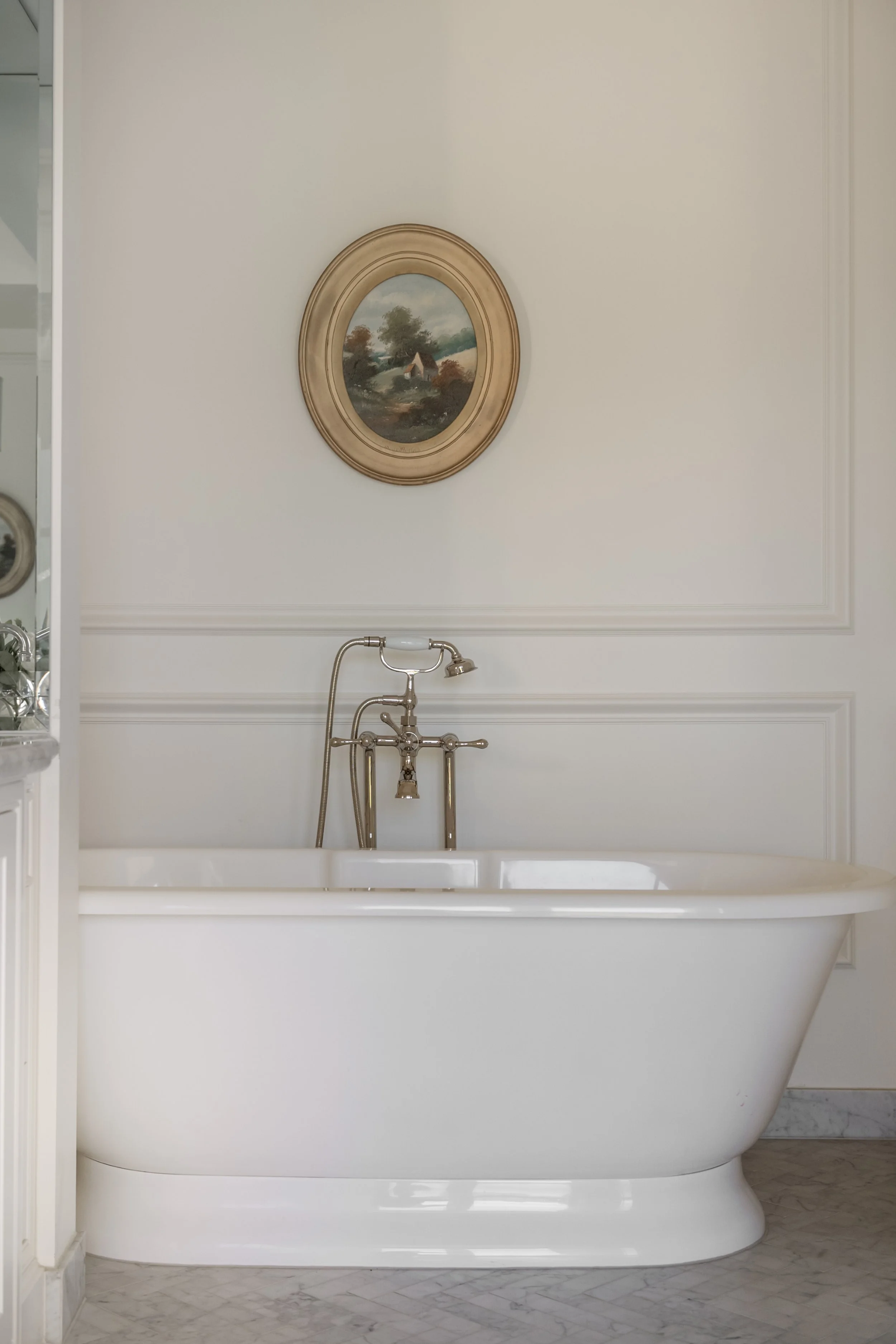 Vintage white bathtub with brass fixtures in a bathroom, framed landscape painting above, marble floor.