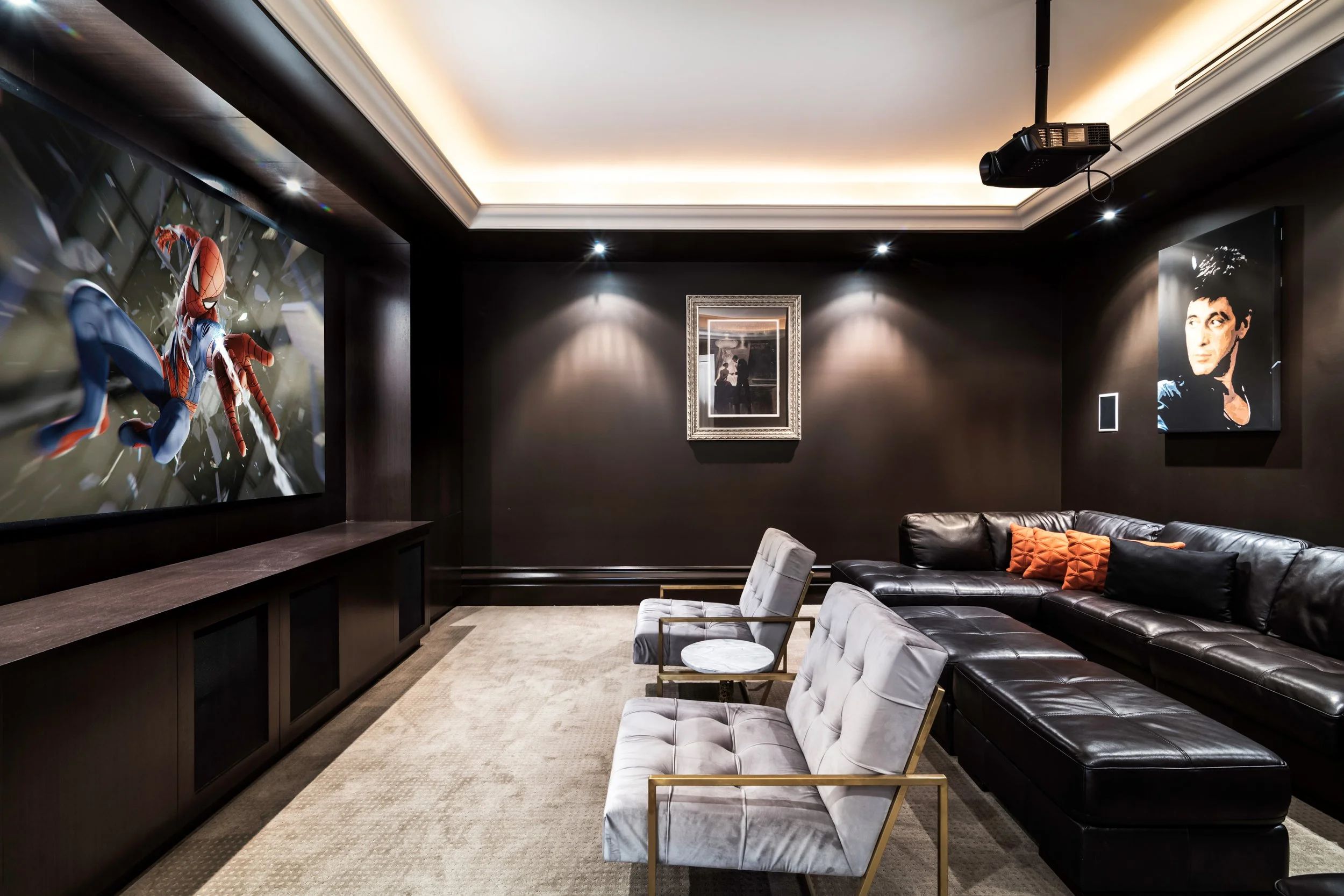 A modern home theater room with a large screen displaying Spider-Man in action, black walls with framed pictures, a beige carpet, a black leather sectional sofa with orange and black pillows, two white armchairs with gold accents, a ceiling-mounted p