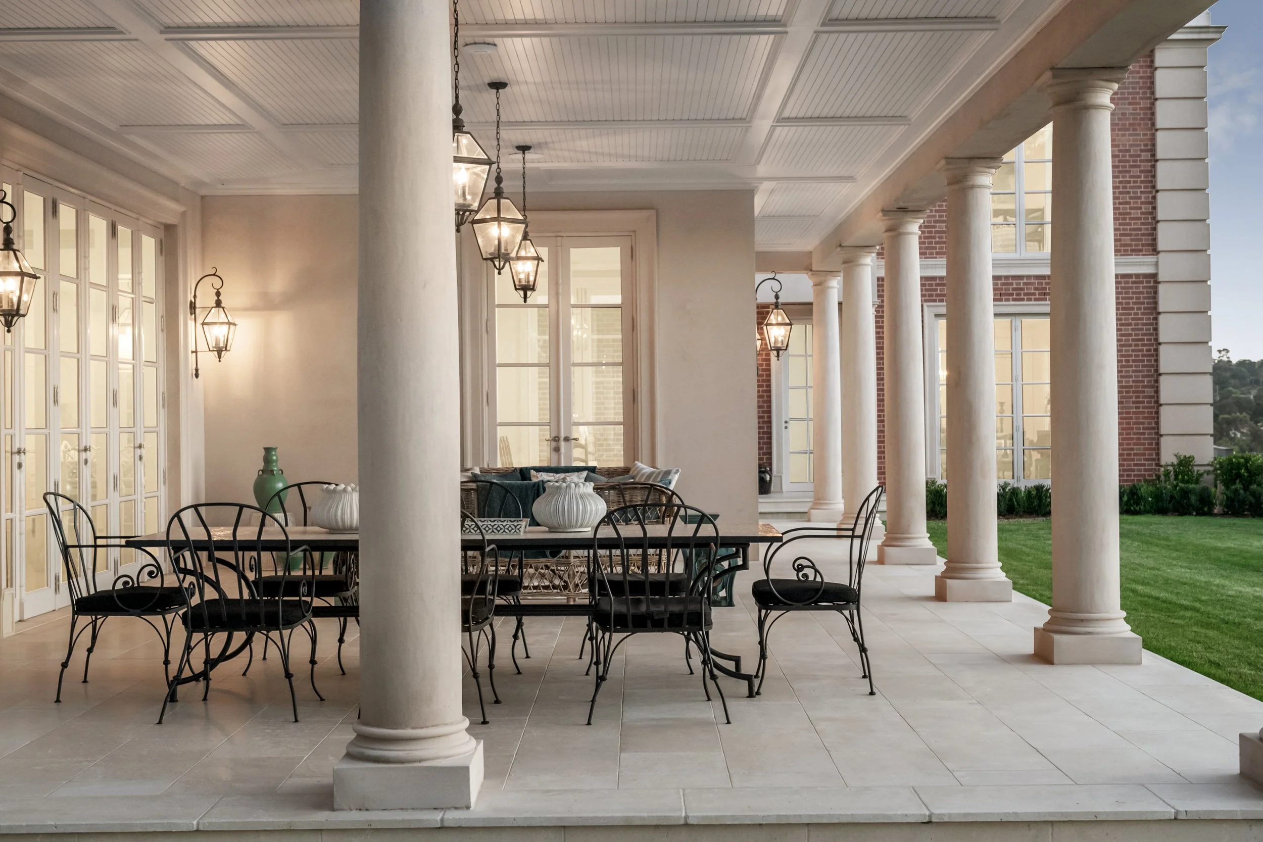 Covered porch with a dining table, black metal chairs, and hanging lantern-style lights, with columns supporting the roof, and a view of a brick building and green lawn outside.