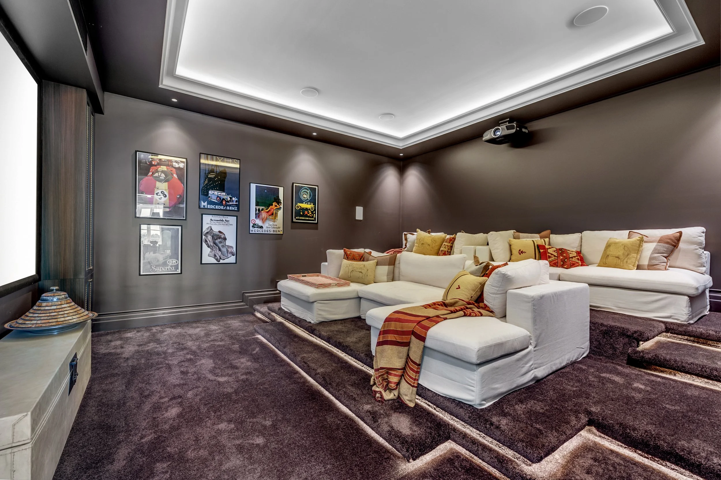 Home theater room with a white sectional sofa, yellow and red cushions, a dark carpet, and framed posters on the wall.