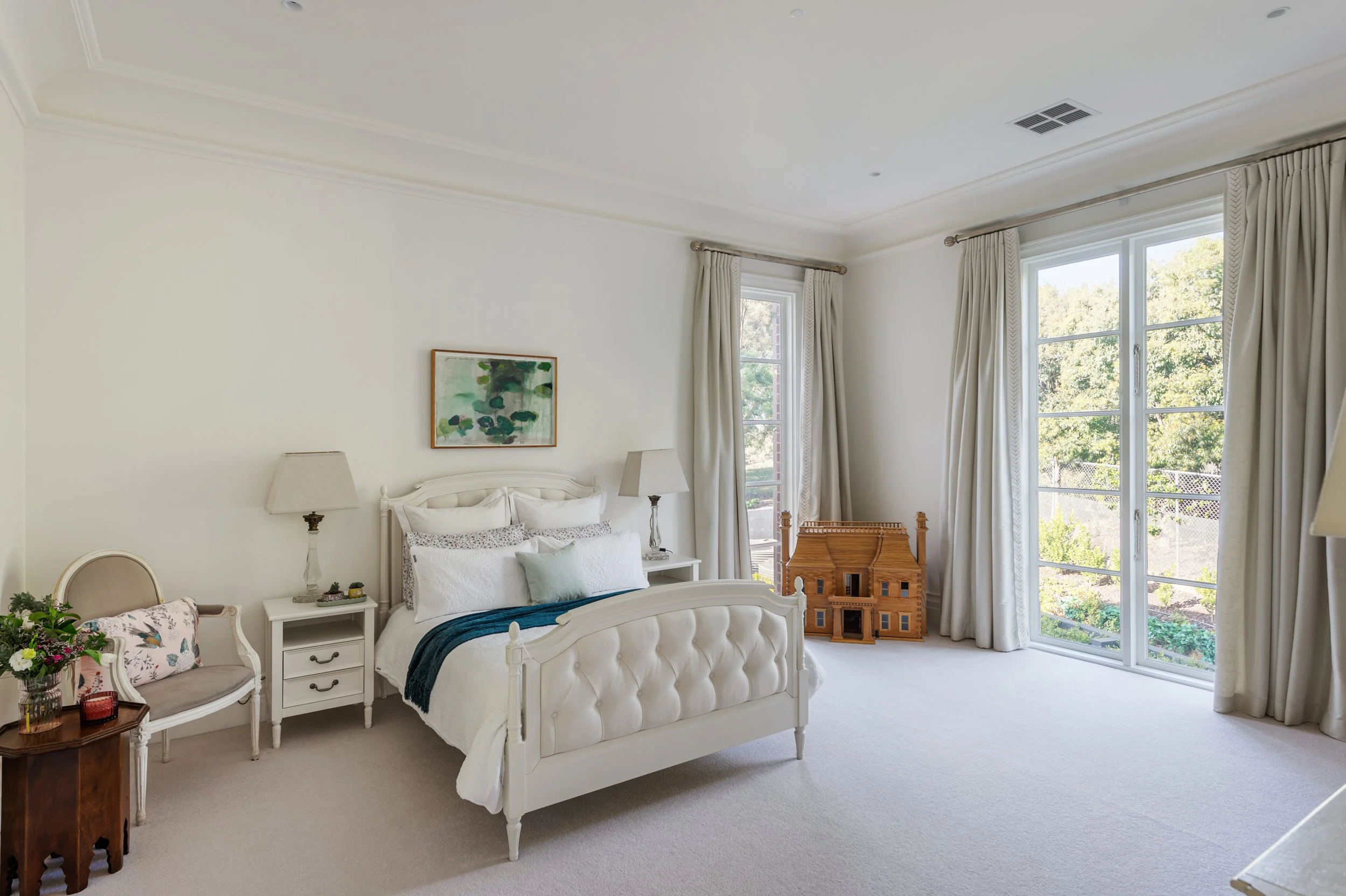 A bright bedroom with a white bed, side tables, lamps, a painting, a small bench with flowers, and a wooden dollhouse near large windows with white curtains and a view of trees.