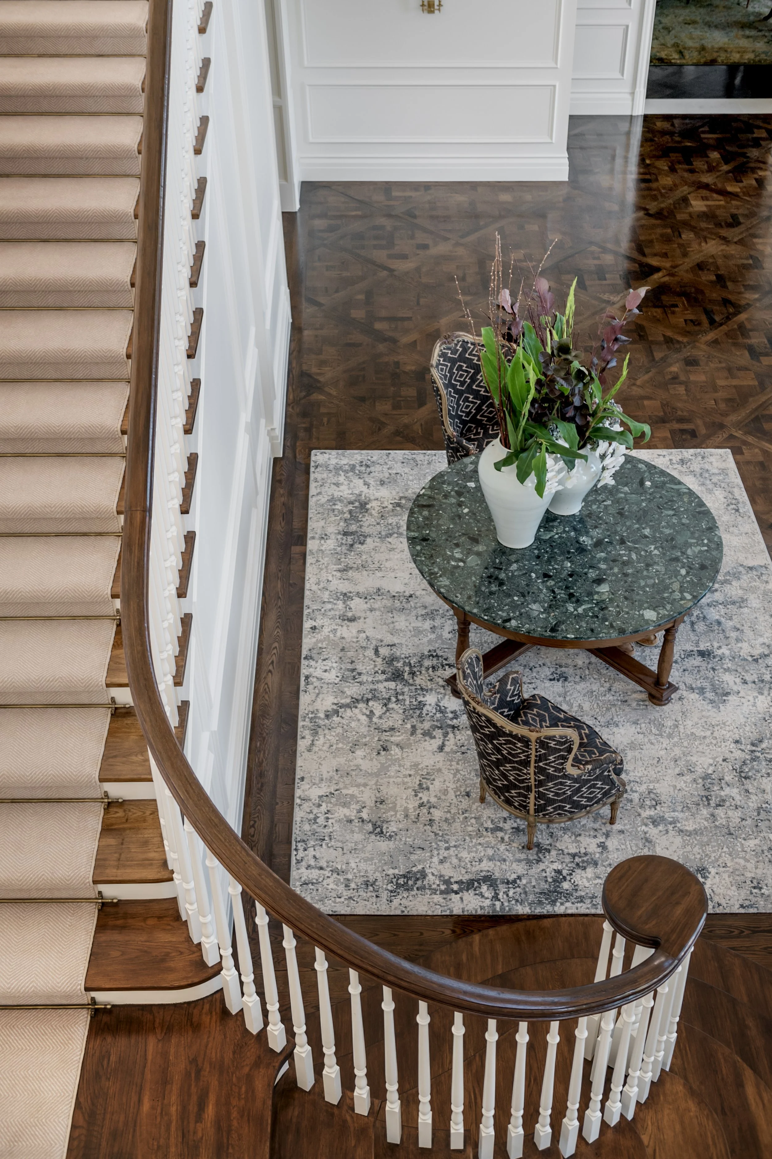 View of a staircase, a small round table with a green marble top, two patterned chairs, a vase with flowers, and a rug on a wooden floor.
