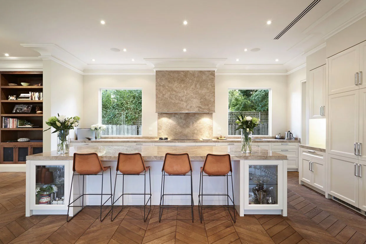Modern kitchen with white cabinets, a large island with barstools, two windows with views of greenery, and a brown tiled floor.