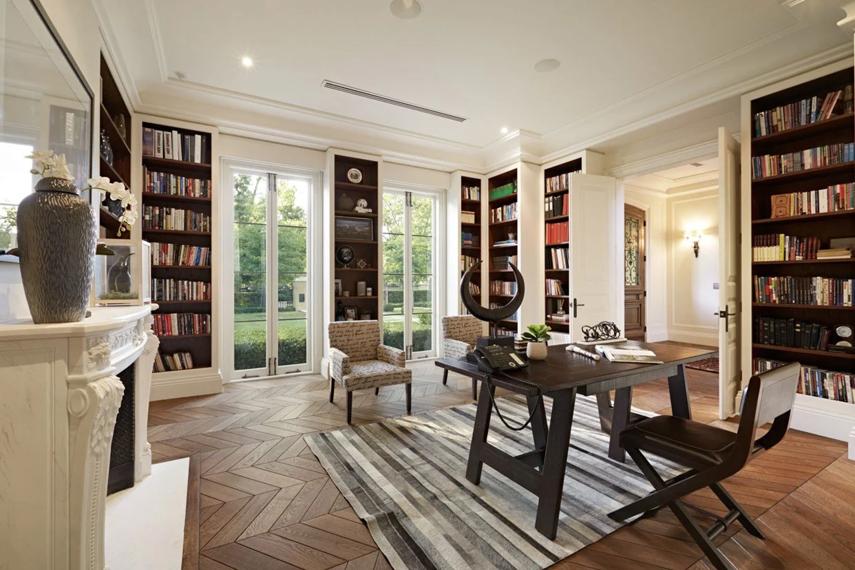 A bright, elegant home office with built-in bookshelves full of books, a wooden desk with papers and a telephone, two patterned chairs, a striped rug, and two large windows overlooking a green yard.