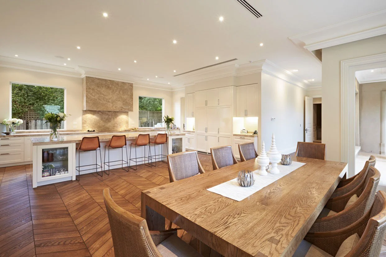 Spacious open-concept dining and kitchen area with wooden dining table, eight chairs, white cabinetry, bar stools, and multiple windows in a bright, modern home.