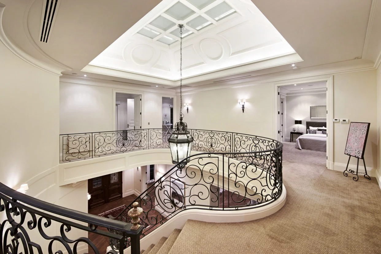 Interior view of a luxurious home or hotel with a curved staircase featuring ornate black wrought iron railings, a hanging lantern, and a lighting ceiling with a skylight, leading to a hallway with open doors revealing rooms.