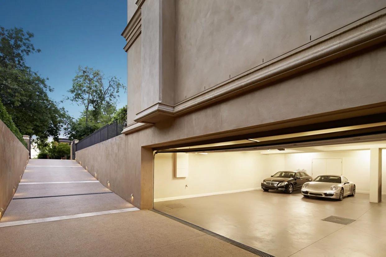 A closed garage with two luxury cars inside, and an upward-sloping driveway outside leading to a residential area with trees.