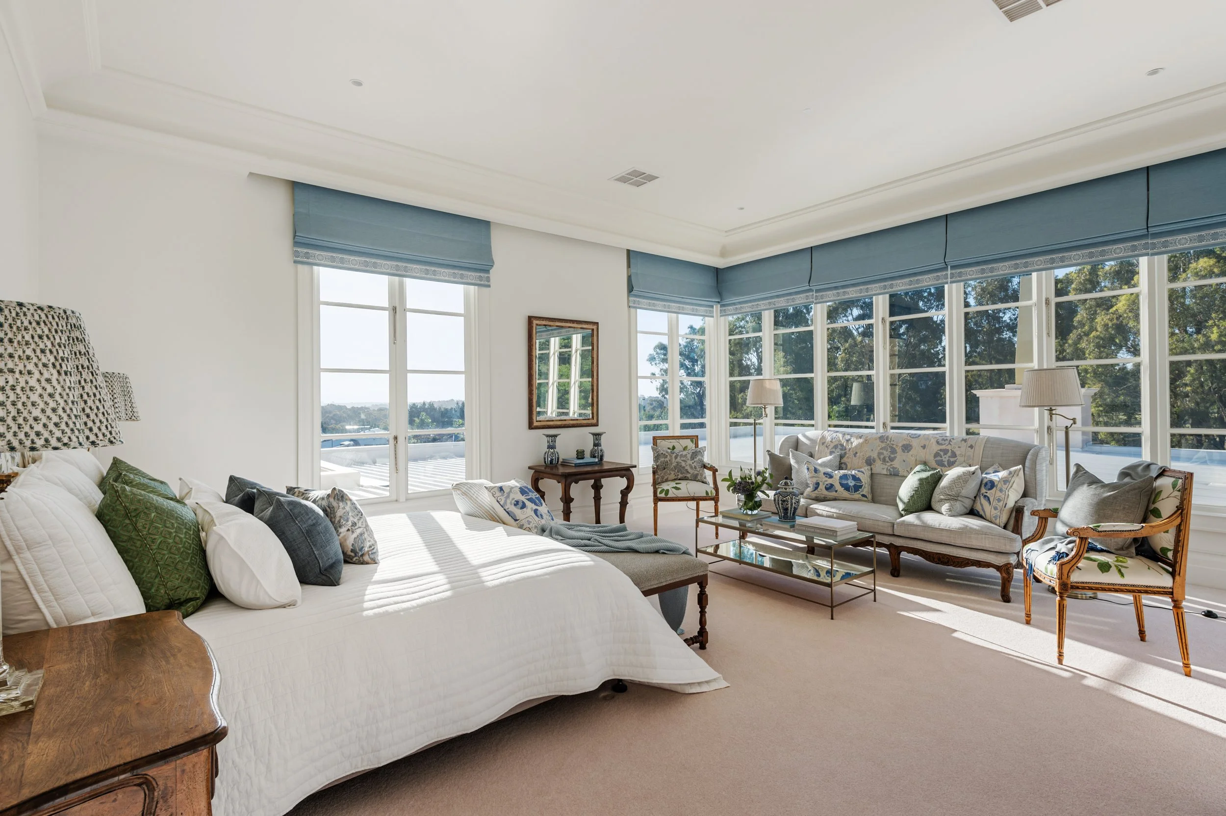 Bright living room with large windows, white walls, and beige carpet; furnished with white sofa, chairs, and wooden side tables; decorated with pillows, lamps, vases, and framed mirror.