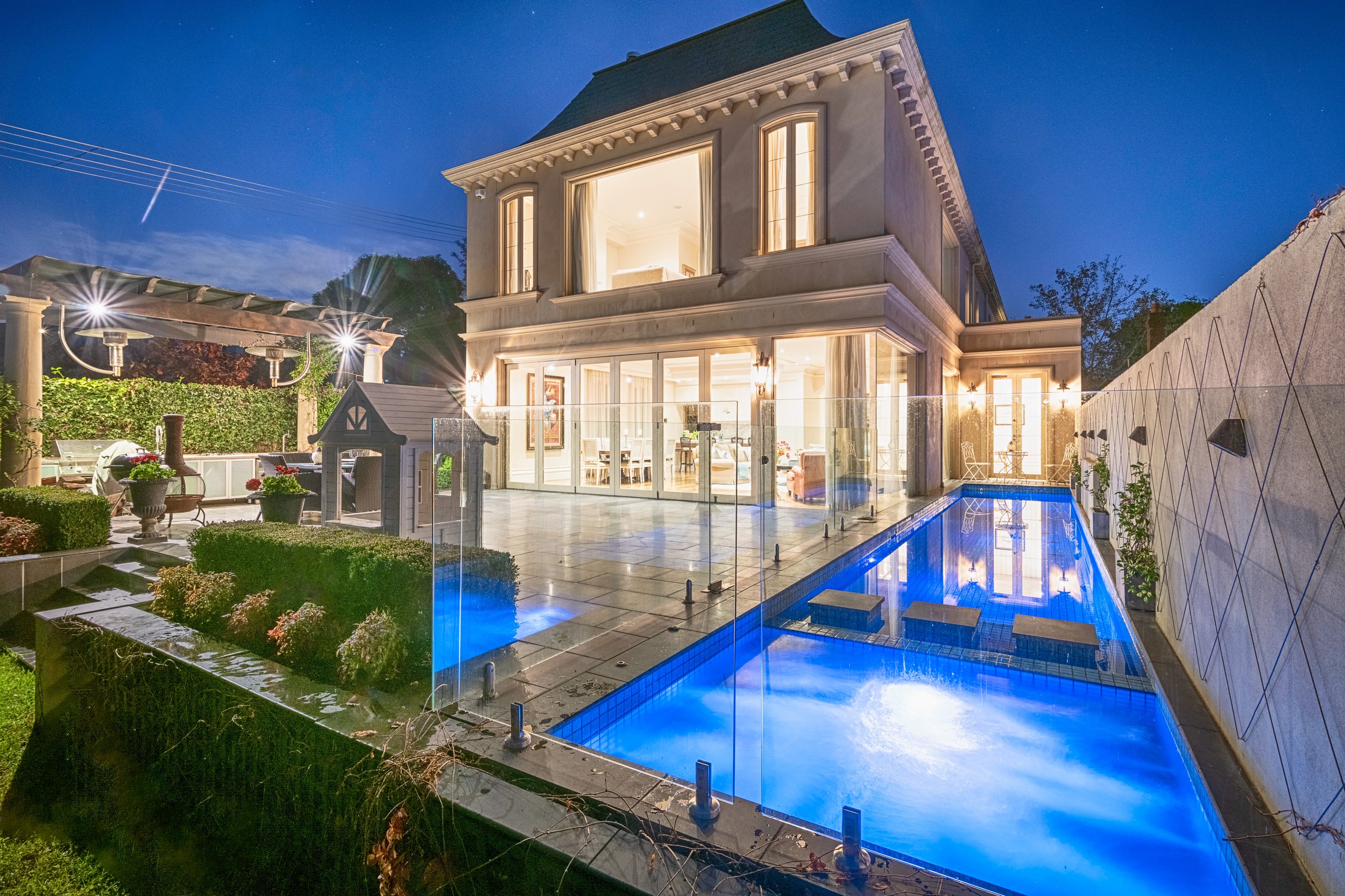 Modern two-story house with large glass windows, illuminated inside, next to a lit swimming pool at night.