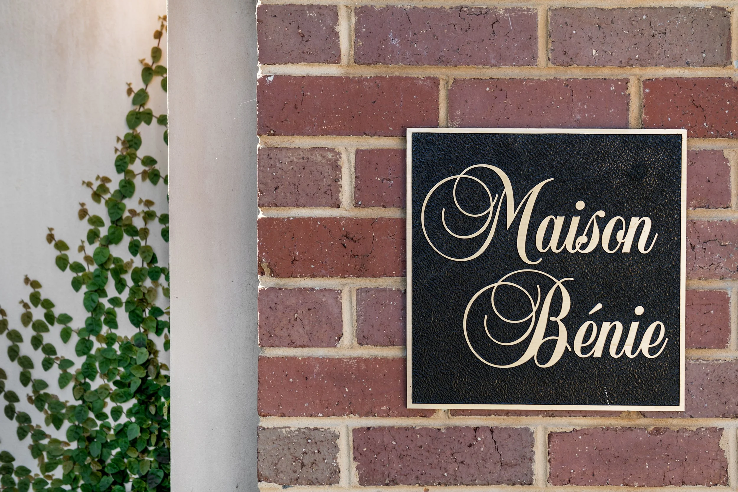 A black decorative sign with the words "Maison Bénie" in elegant script on a brick wall, with green ivy on the side.