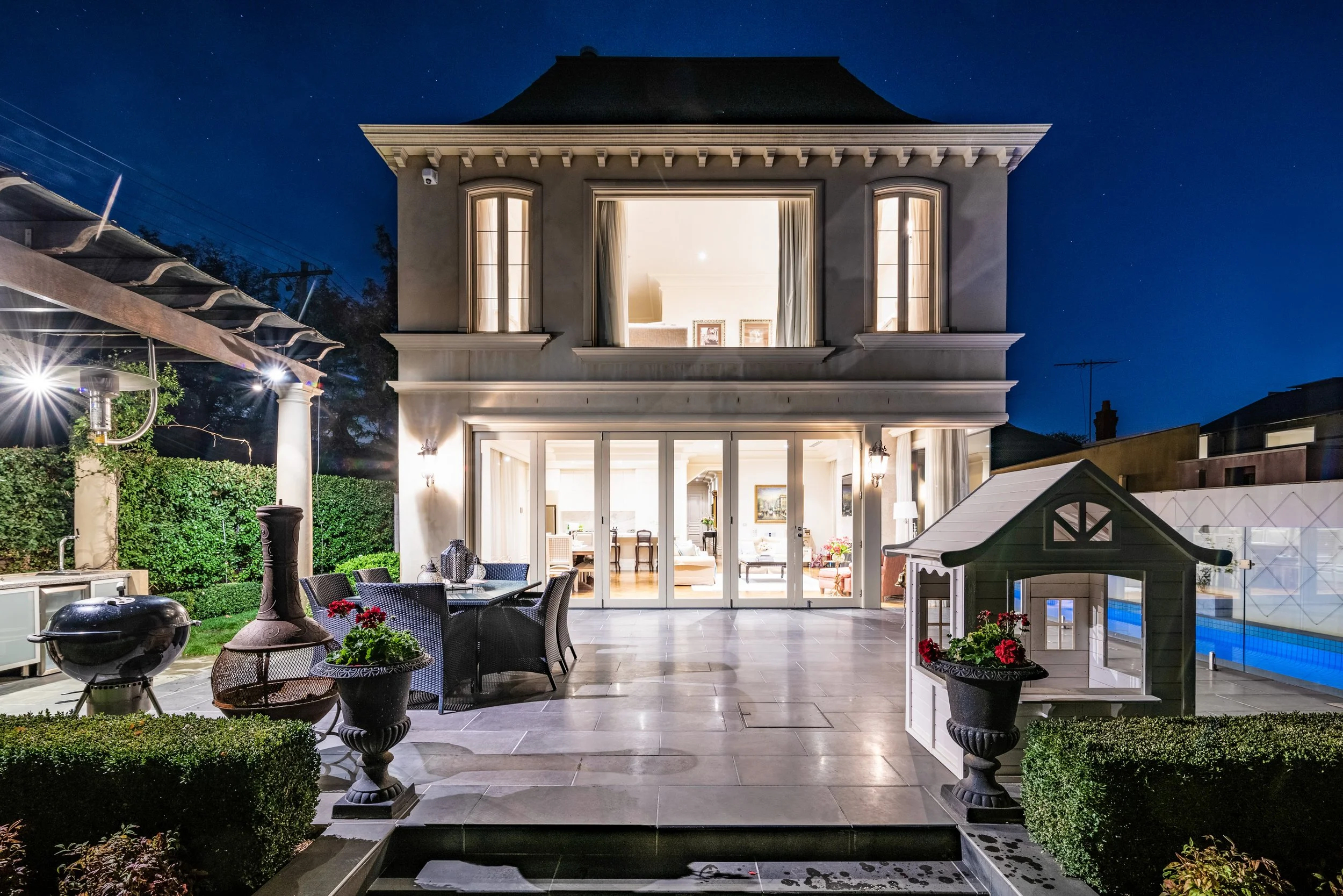Night view of a modern house with large glass doors leading to a patio, illuminated from within. The patio has outdoor furniture, potted plants, a barbecue grill, and a small decorative playhouse, all surrounded by greenery.