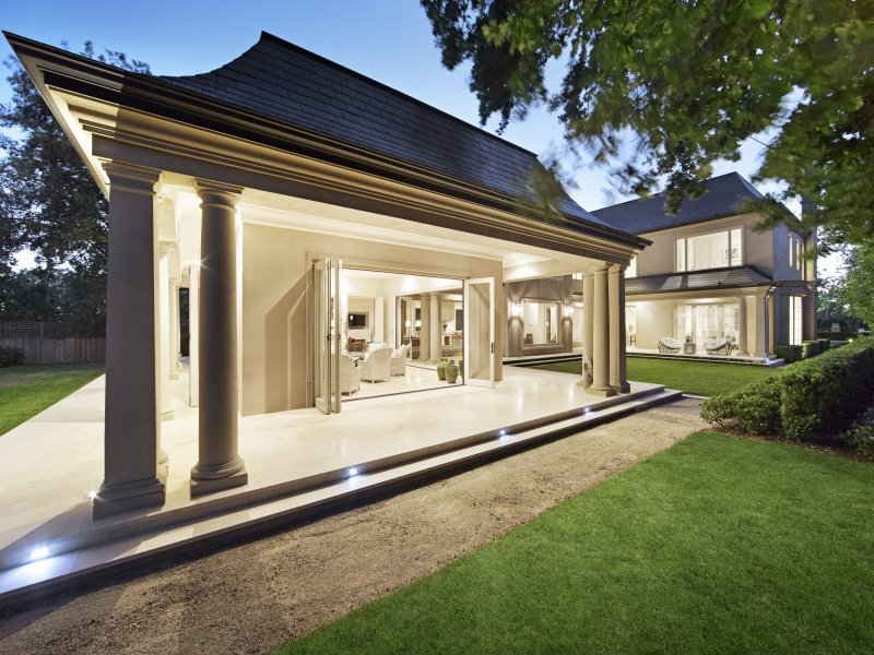 Backyard view of a luxurious house with an open patio area and outdoor seating, illuminated at dusk, with well-maintained lawn and trees.