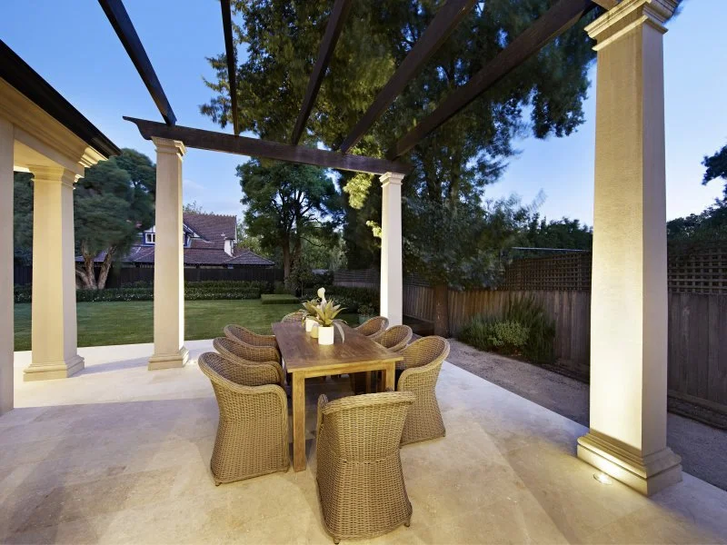Outdoor patio with a wooden dining table and six wicker chairs, decorated with a potted plant, under a pergola with four white columns, surrounded by a green backyard and wooden fence, during evening.