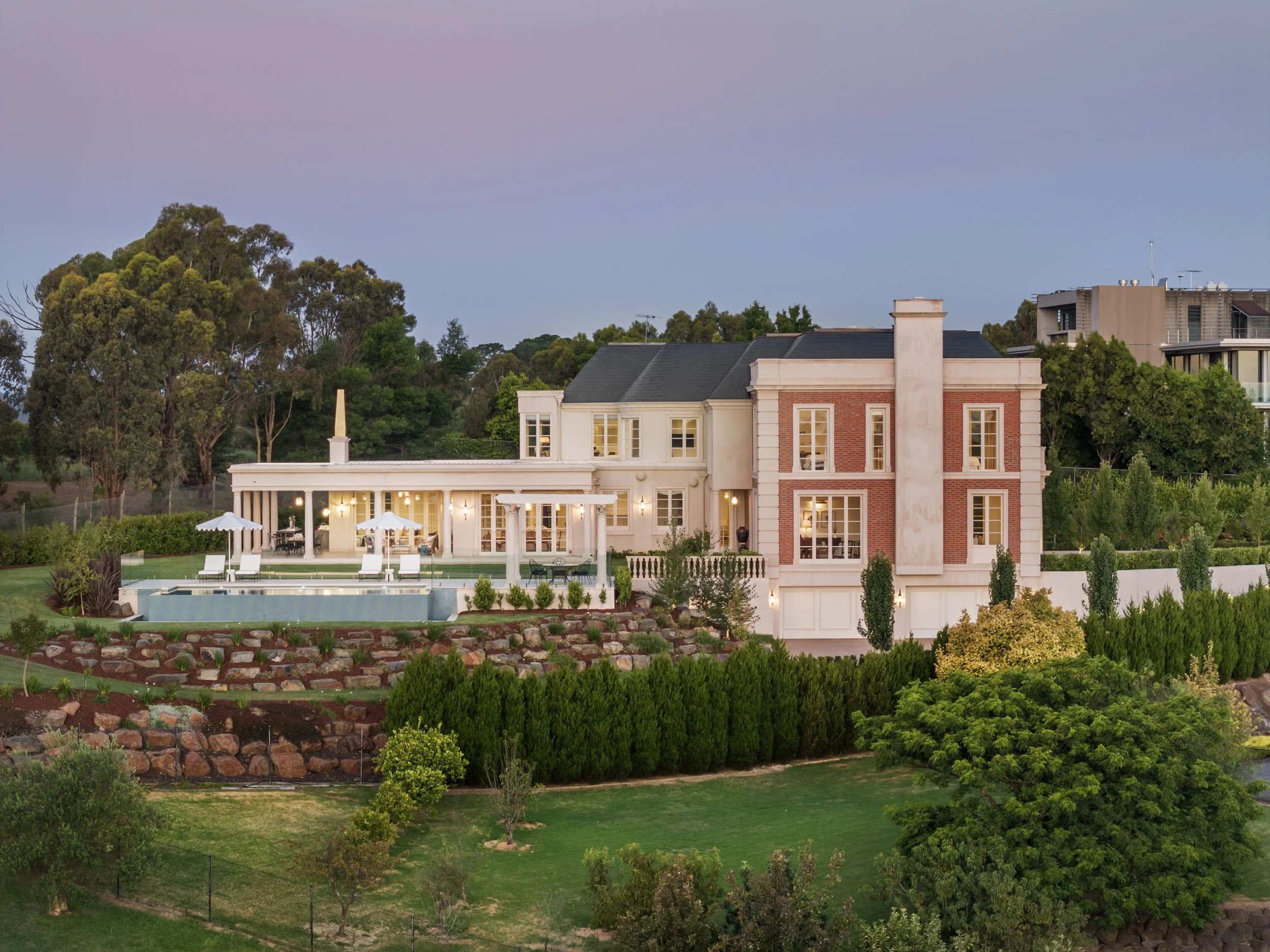 Large mansion with white and brick exterior, outdoor pool area with loungers and umbrellas, surrounded by landscaped garden and trees, during evening with a pinkish sky.