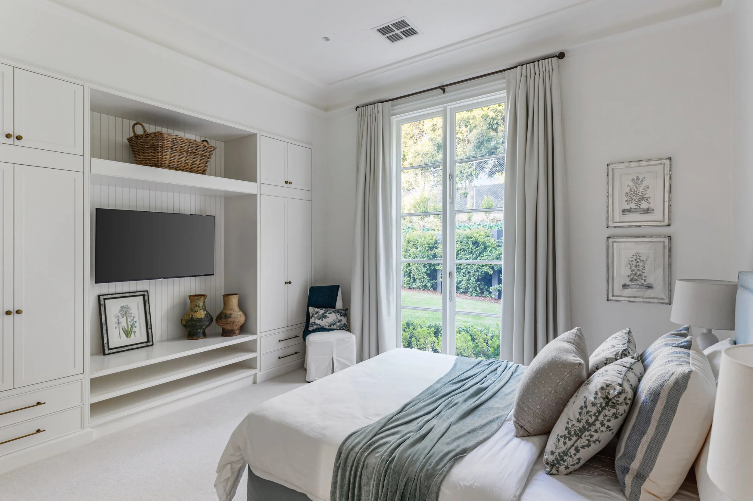 Bright bedroom with white walls, large window with cream curtains, and built-in white wardrobe with open shelves and cabinets. A bed with neutral pillows and a throw blanket is positioned near the window, with artwork on the wall and decorative objec
