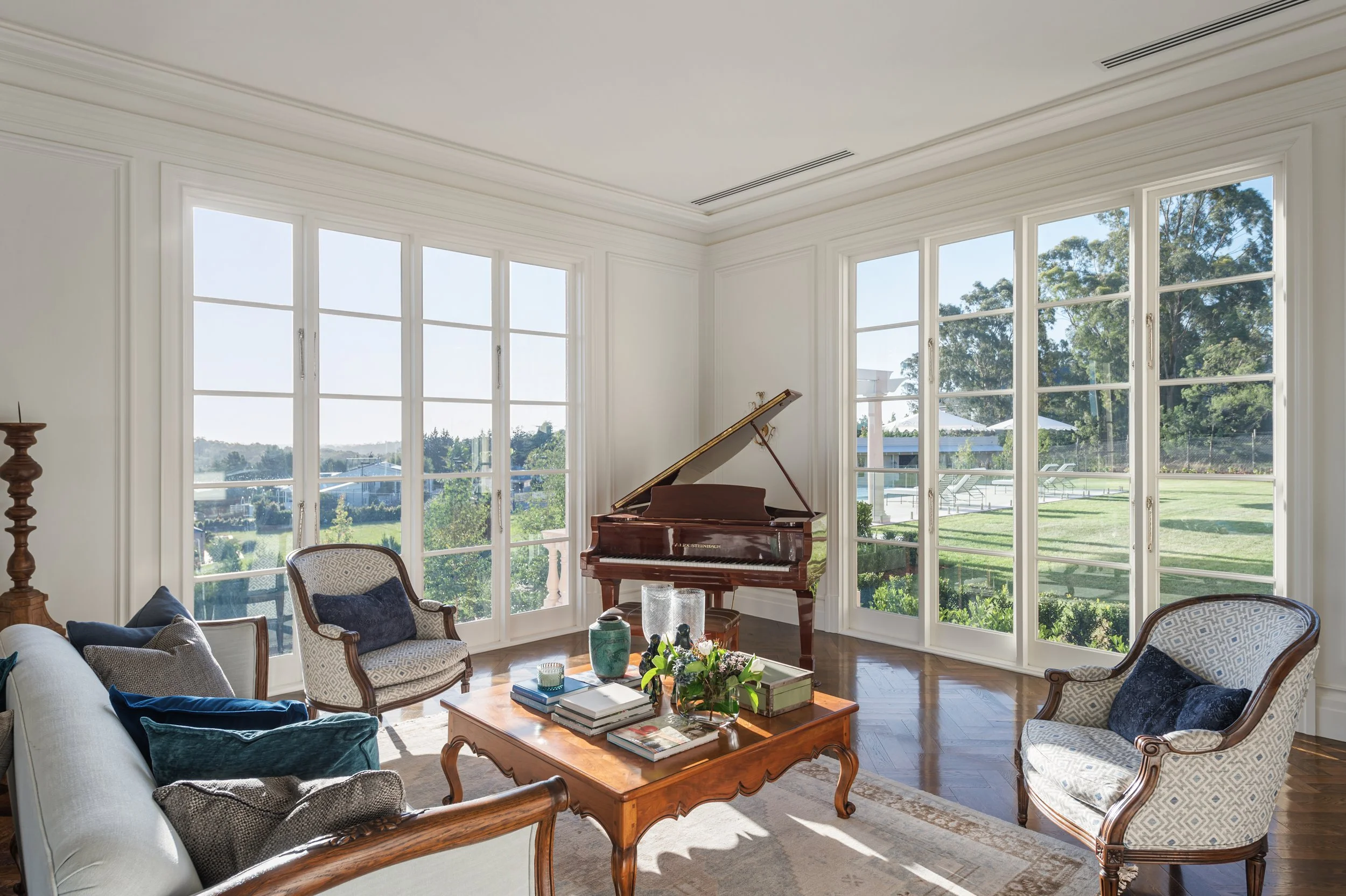 Living room with large windows, a grand piano, a wooden coffee table with books and flowers, and upholstered chairs.